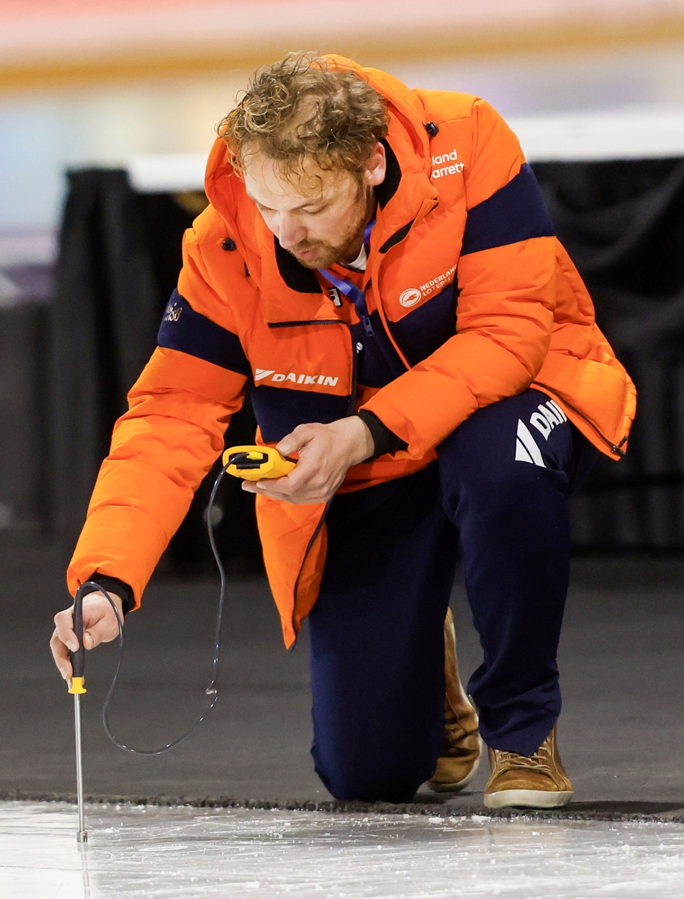 A man in an orange coat kneels down and holds a tool up to ice