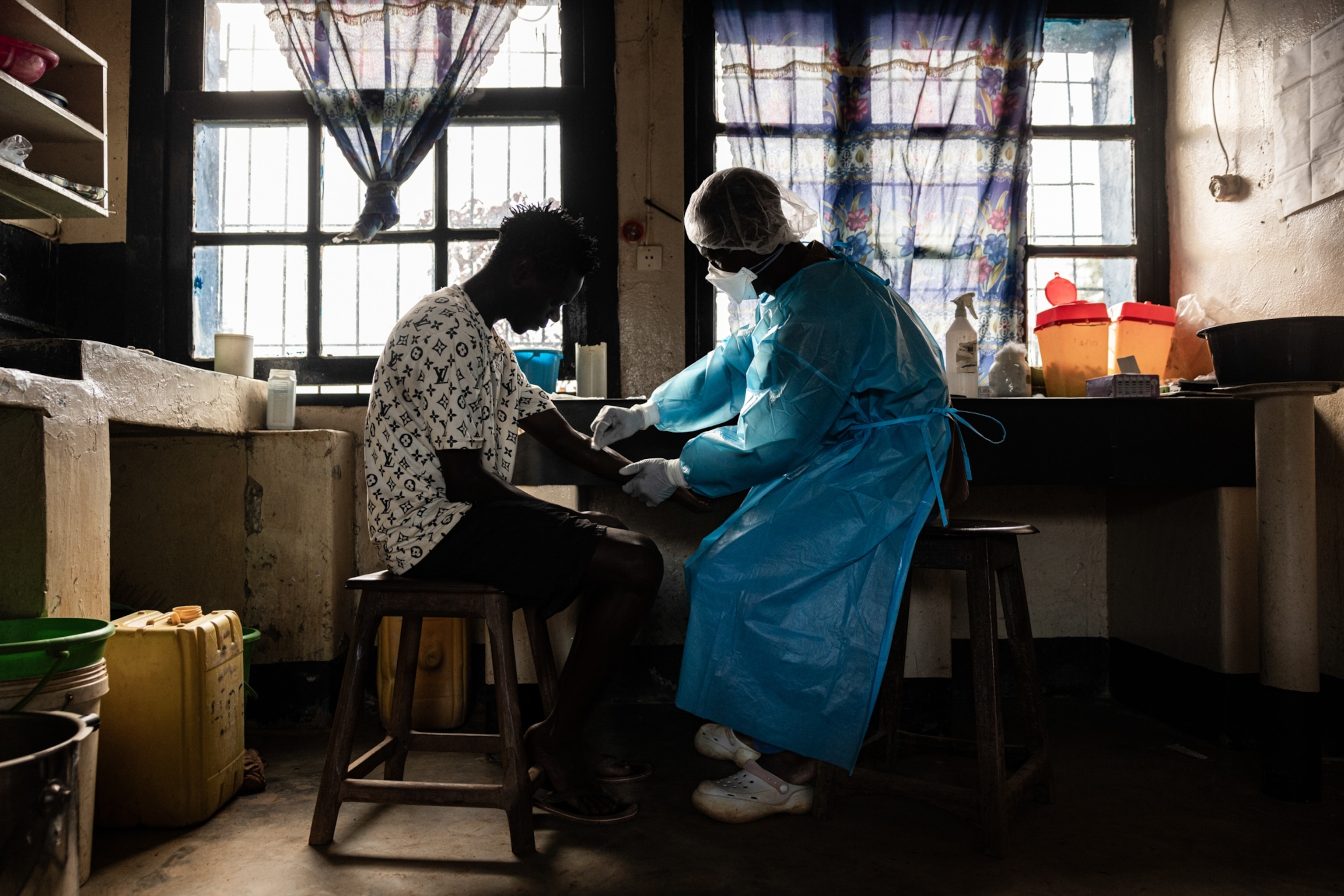 A healthcare worker in protective gear attends to a seated patient in a dimly lit room