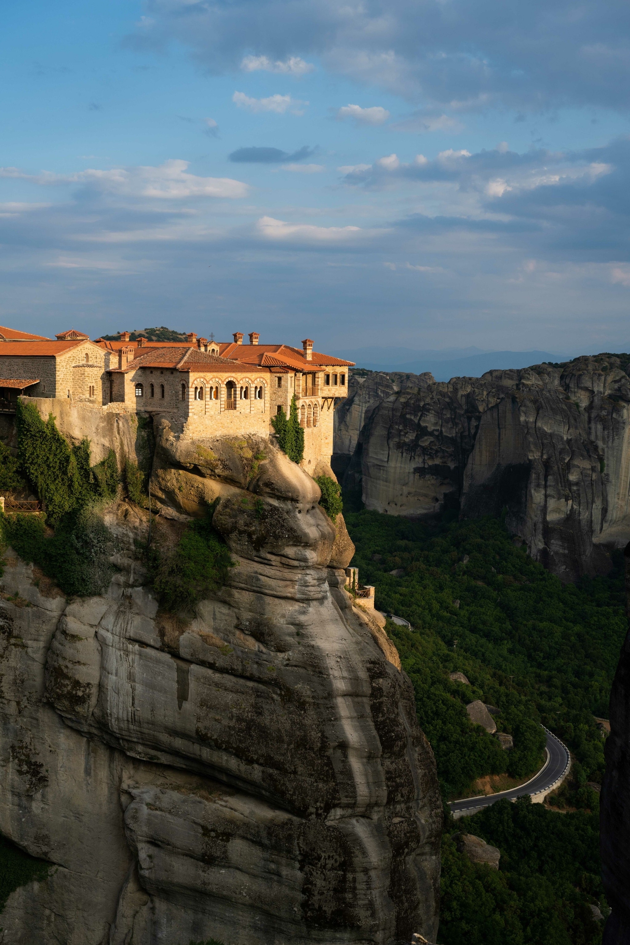 The towering monasteries of Meteora, caught in the sun. They spill over a steep cliff. A road winds around the bottom far below.