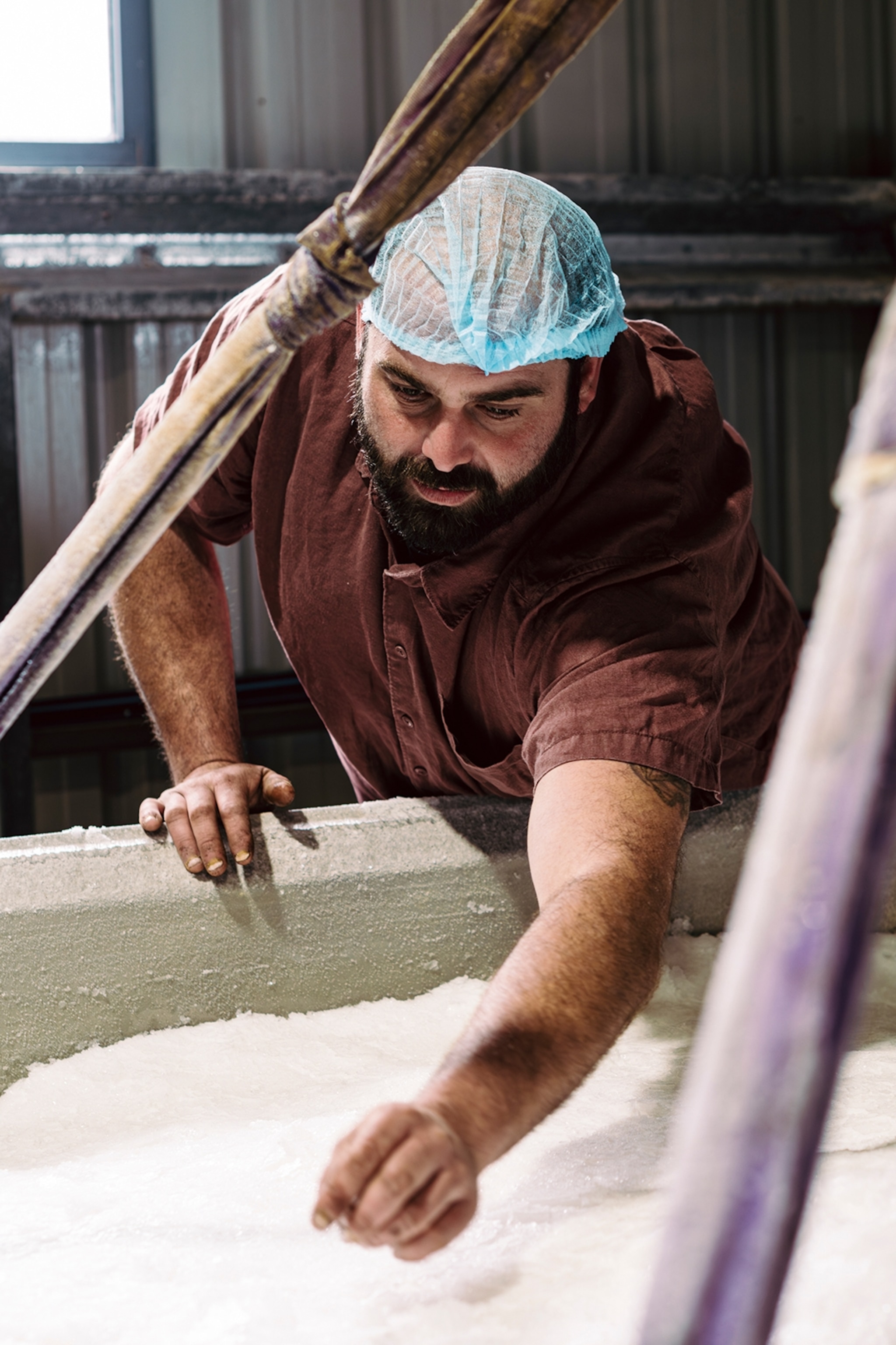 A bearded man wearing a fish net over his hair, checking a salt pan in a factory.
