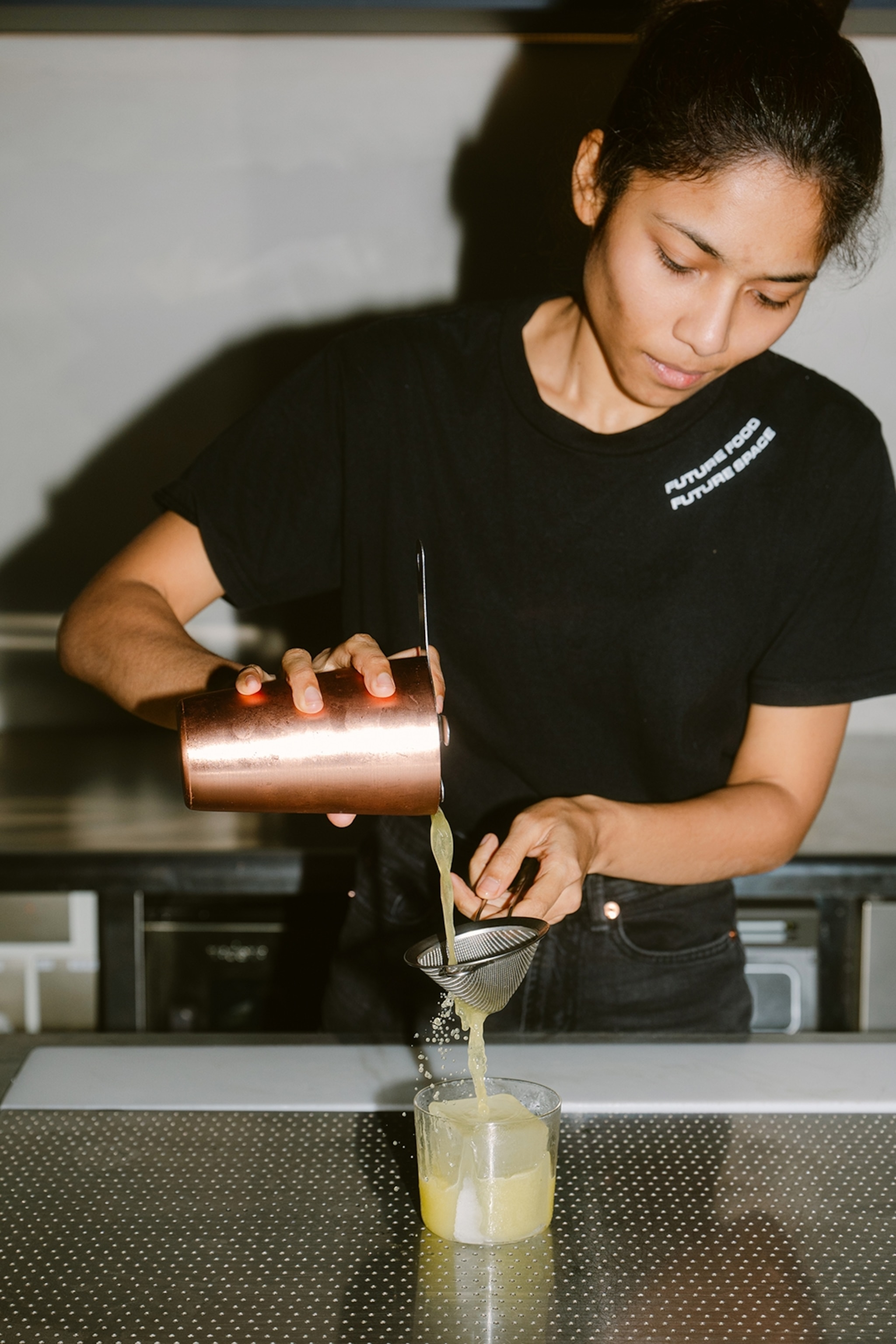 A young, female bartender pouring an opaque drink from a cocktail shaker through a sieve and into a glass tumbler.