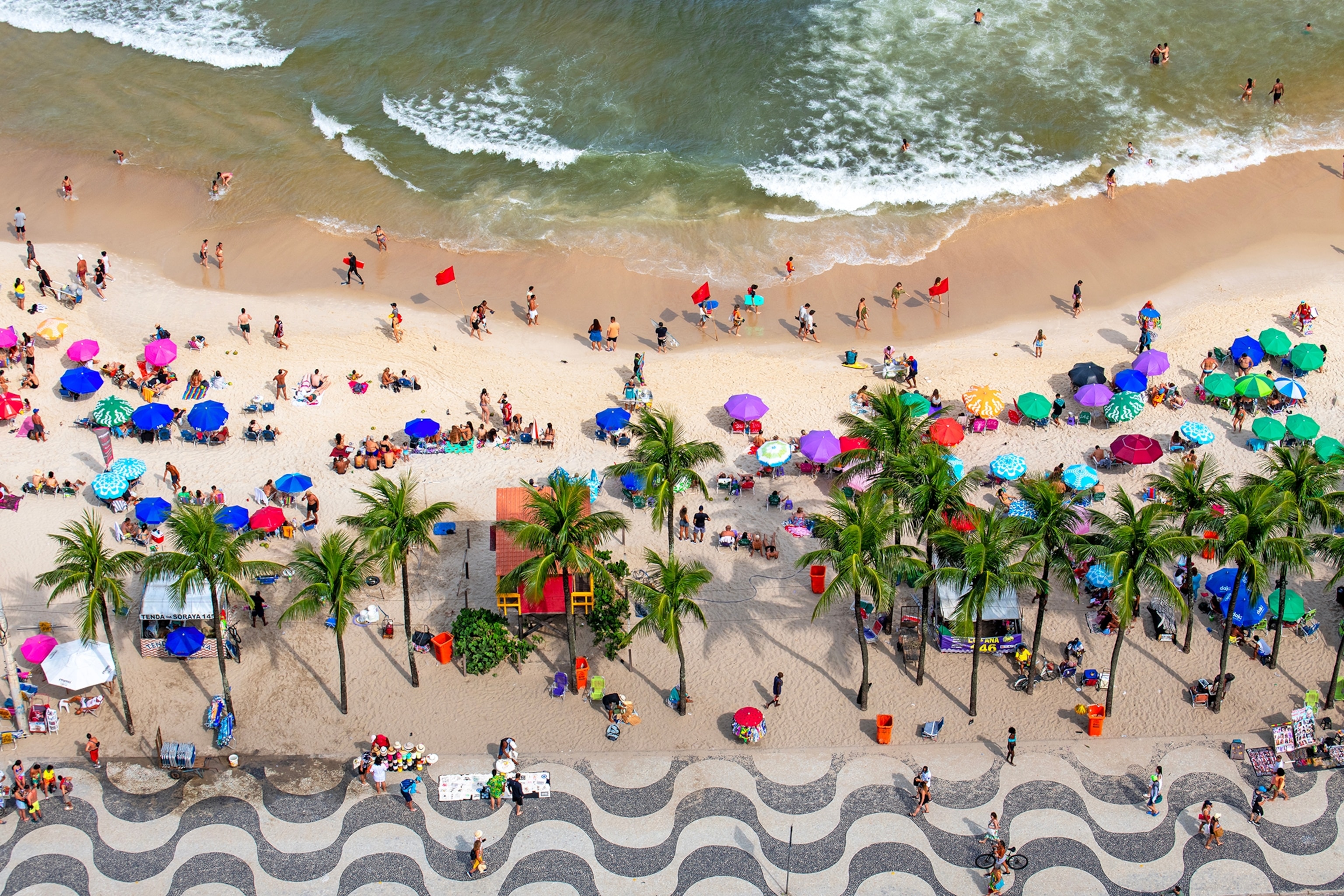 An aerial shot of a beachfront with countless parasols, bathing visitors and a line of palm trees separating the beach and the mosaic sidewalk.