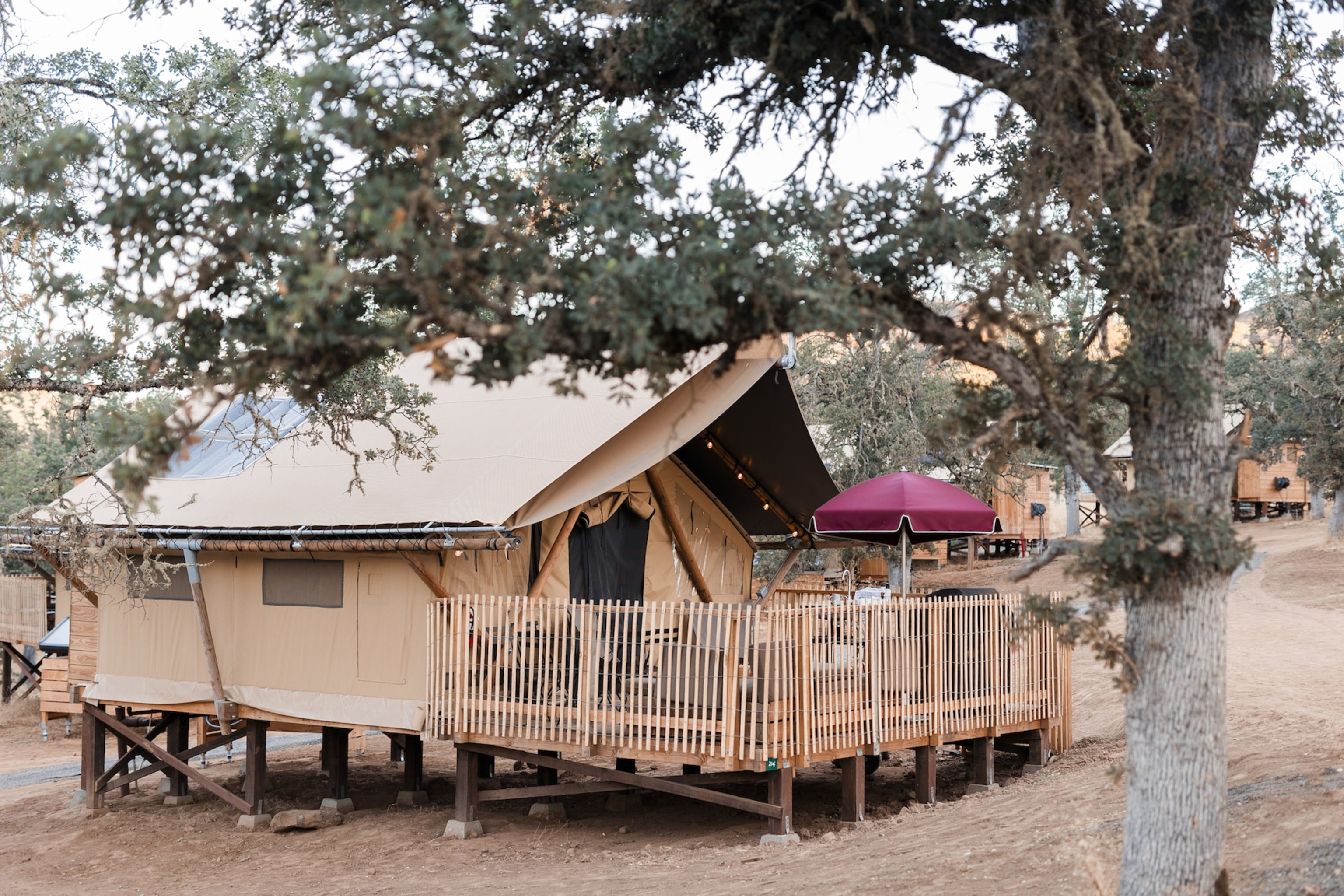 A tan canvas cabin on short lifts with a ramp, a red umbrella on a small deck, and shady trees surrounding