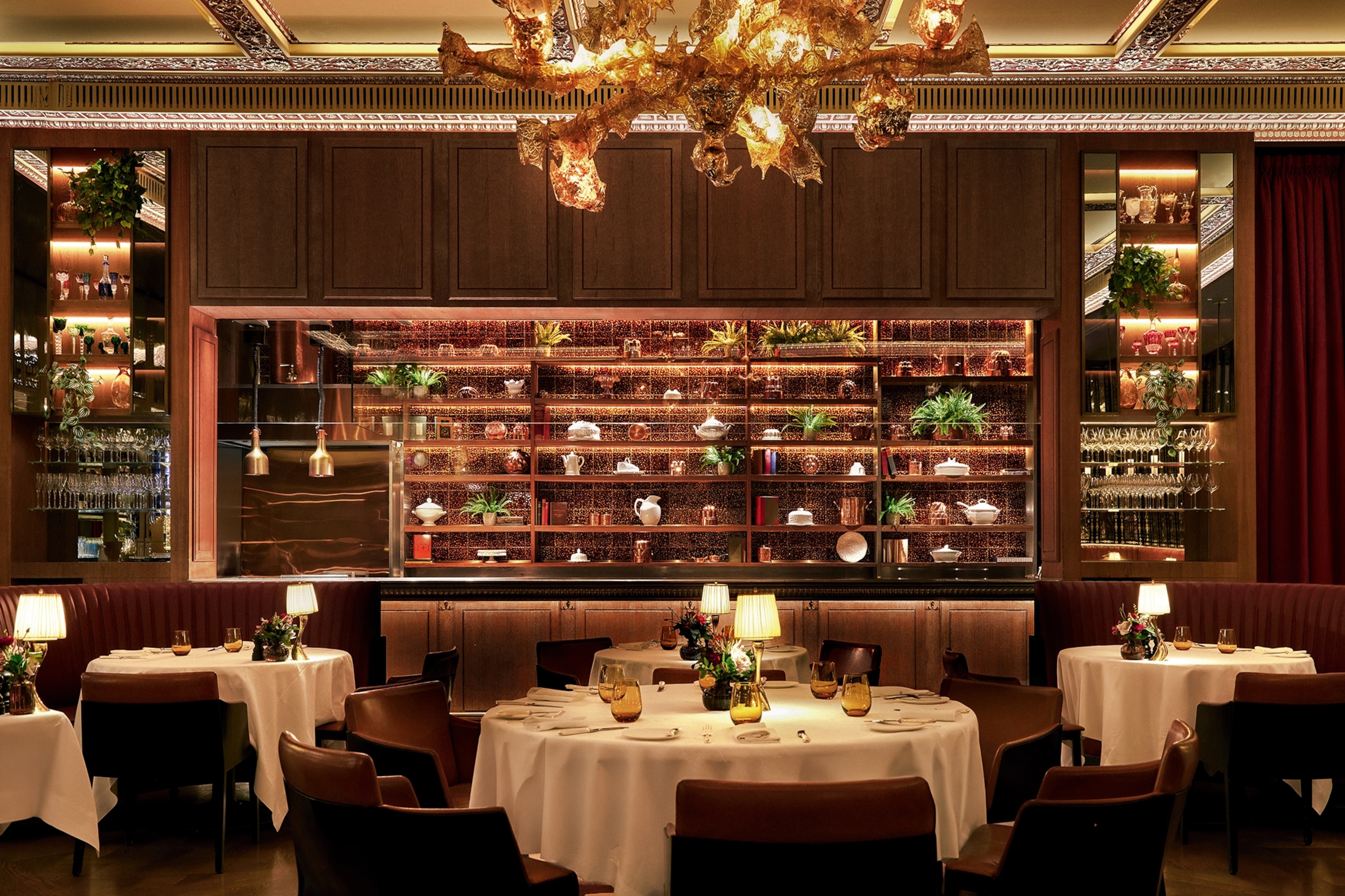 A warmly-lit restaurant dining room with table linen, leather booth seats and a wide, open bar.
