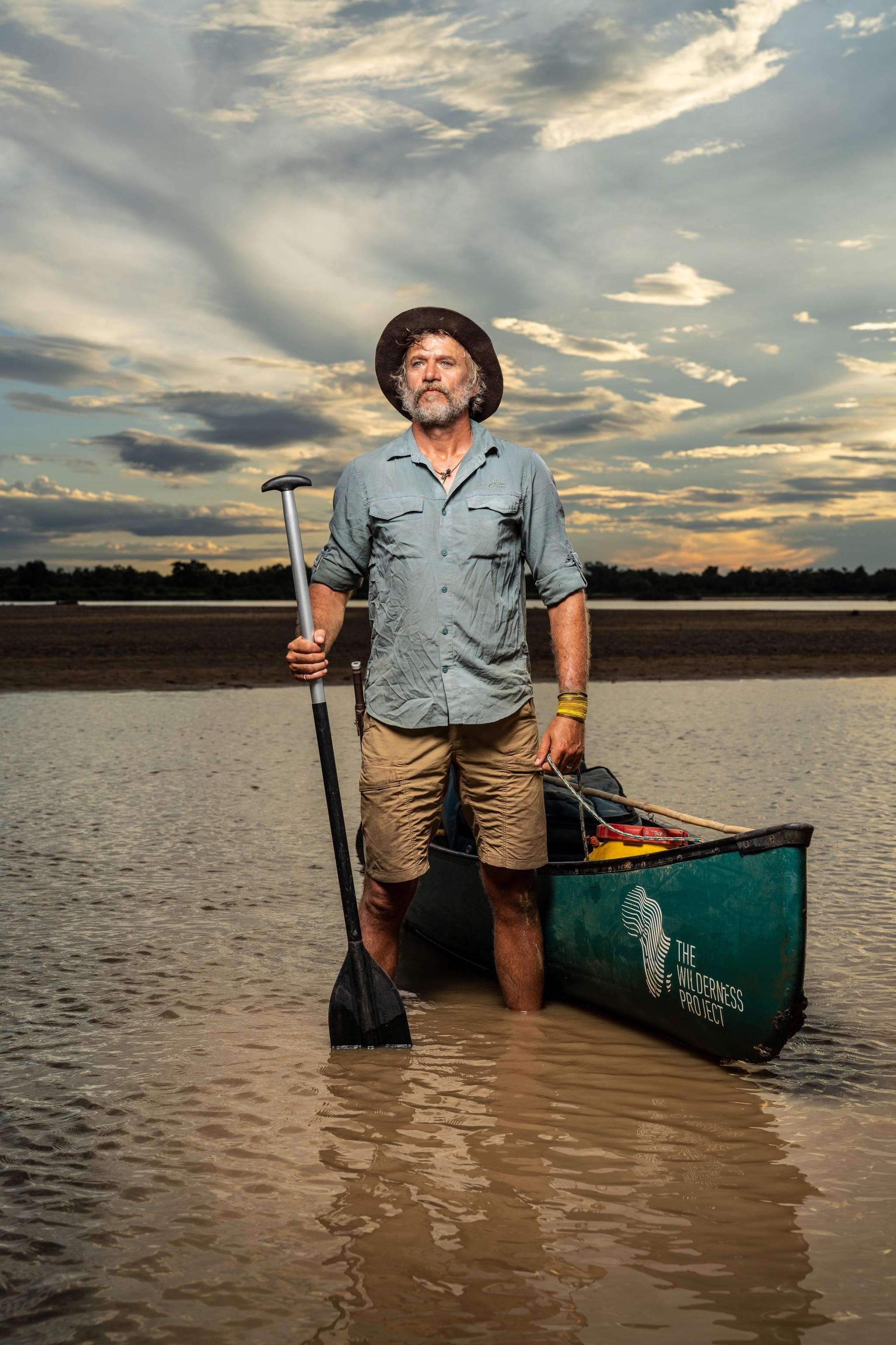 man wearing a hat holding a paddle standing in water next to a green canoe