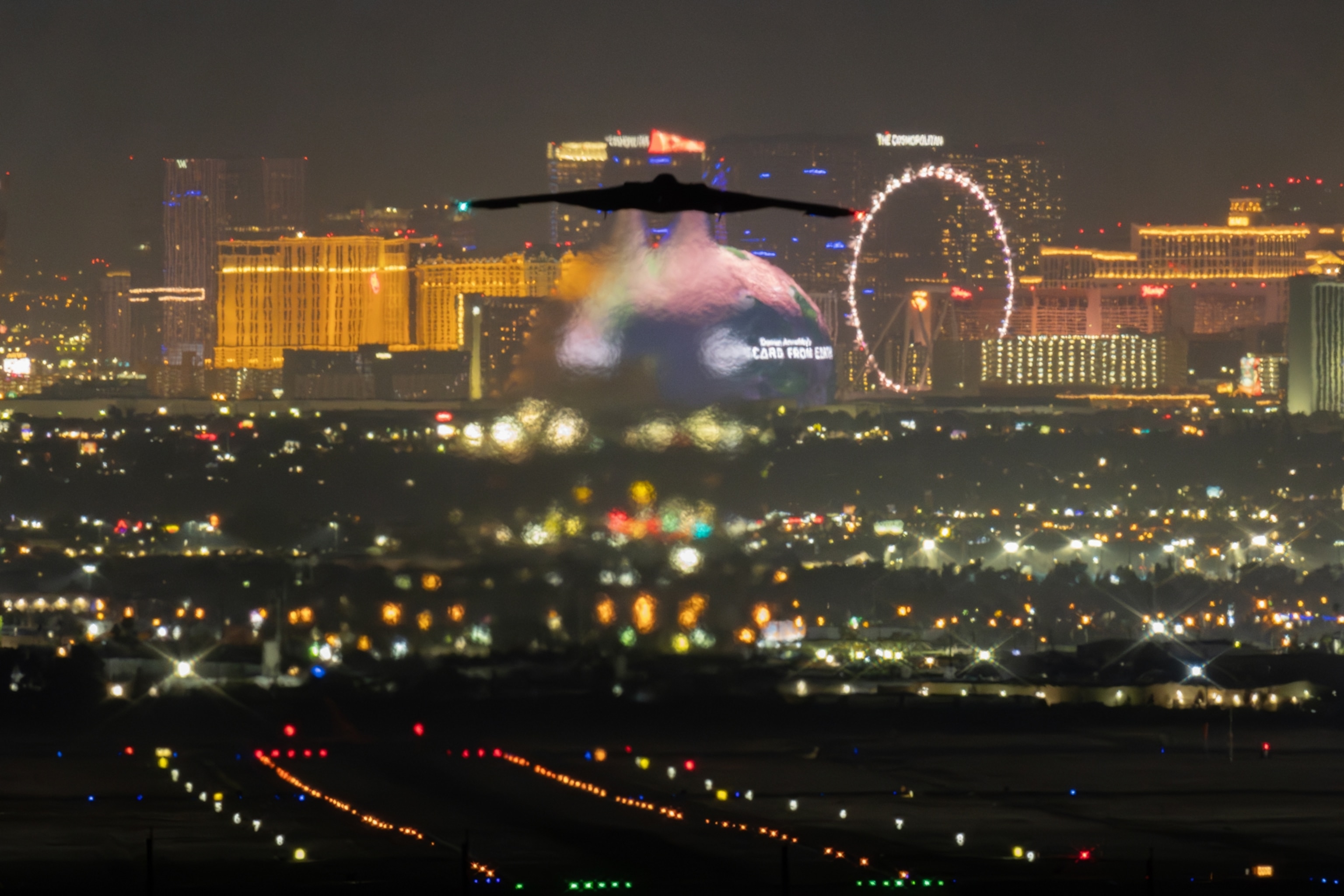 A silhouetted stealth bomber flies over a vibrant cityscape of illuminated buildings and a Ferris wheel in the background