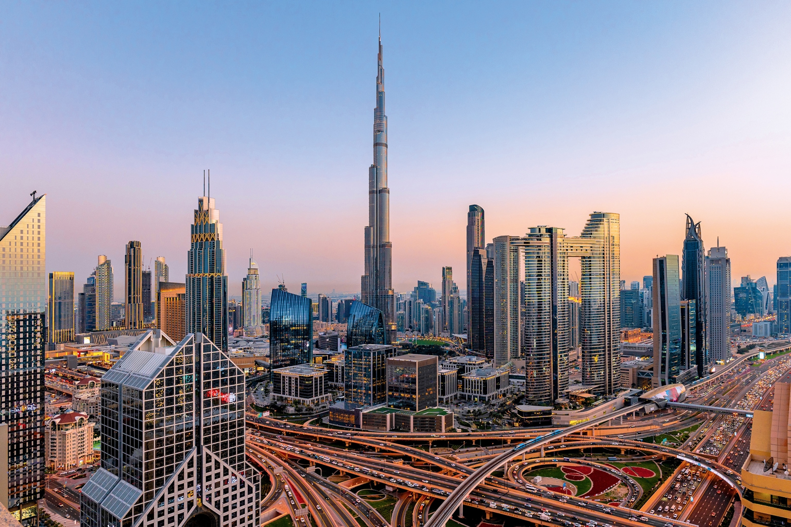 An urban skyline at late dusk, featuring high-reaching, uber-modern skyscrapers and a lit up highway knot.