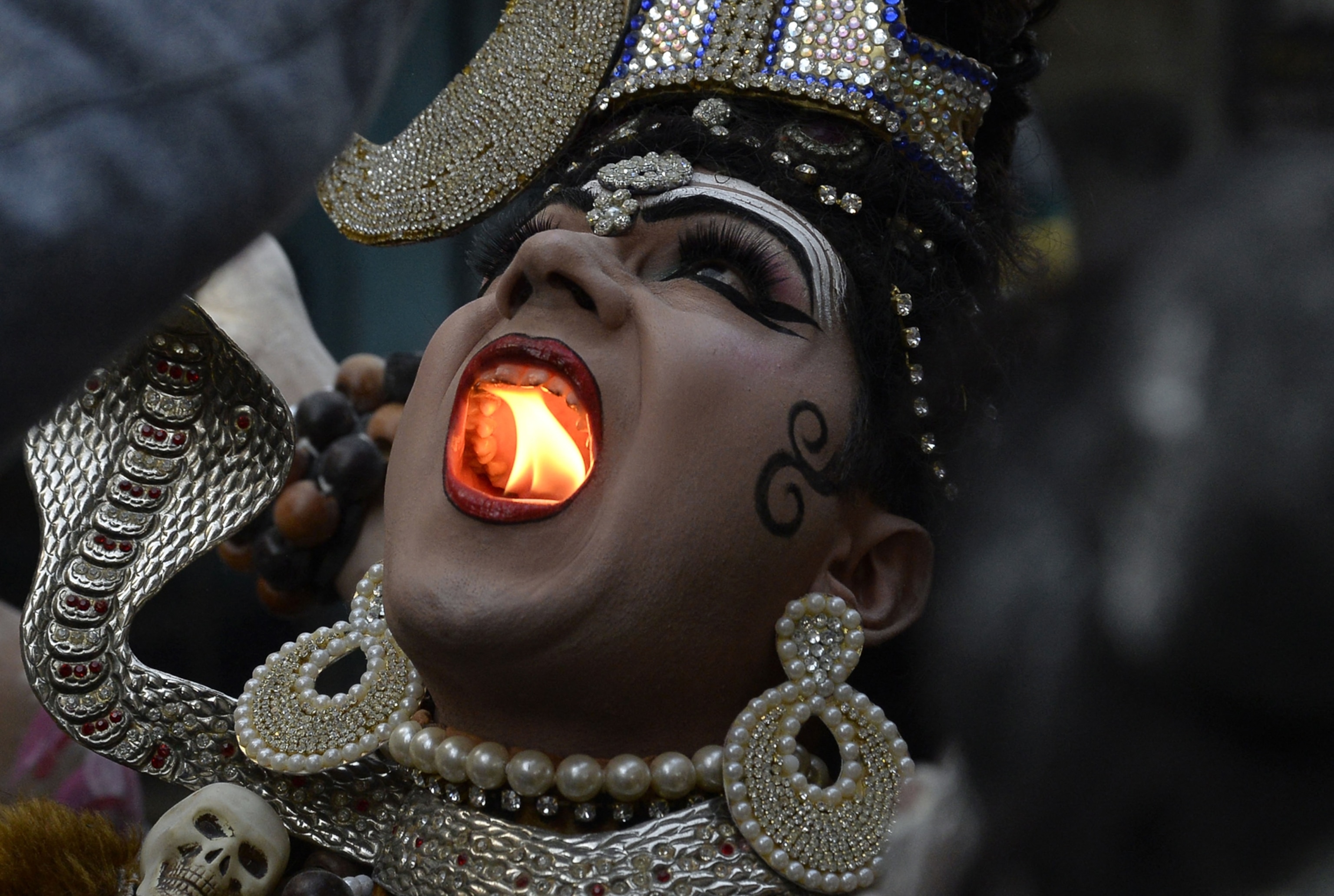 A person dressed in elaborate traditional attire holds a lit object in their open mouth, displaying vibrant jewelry and dramatic makeup
