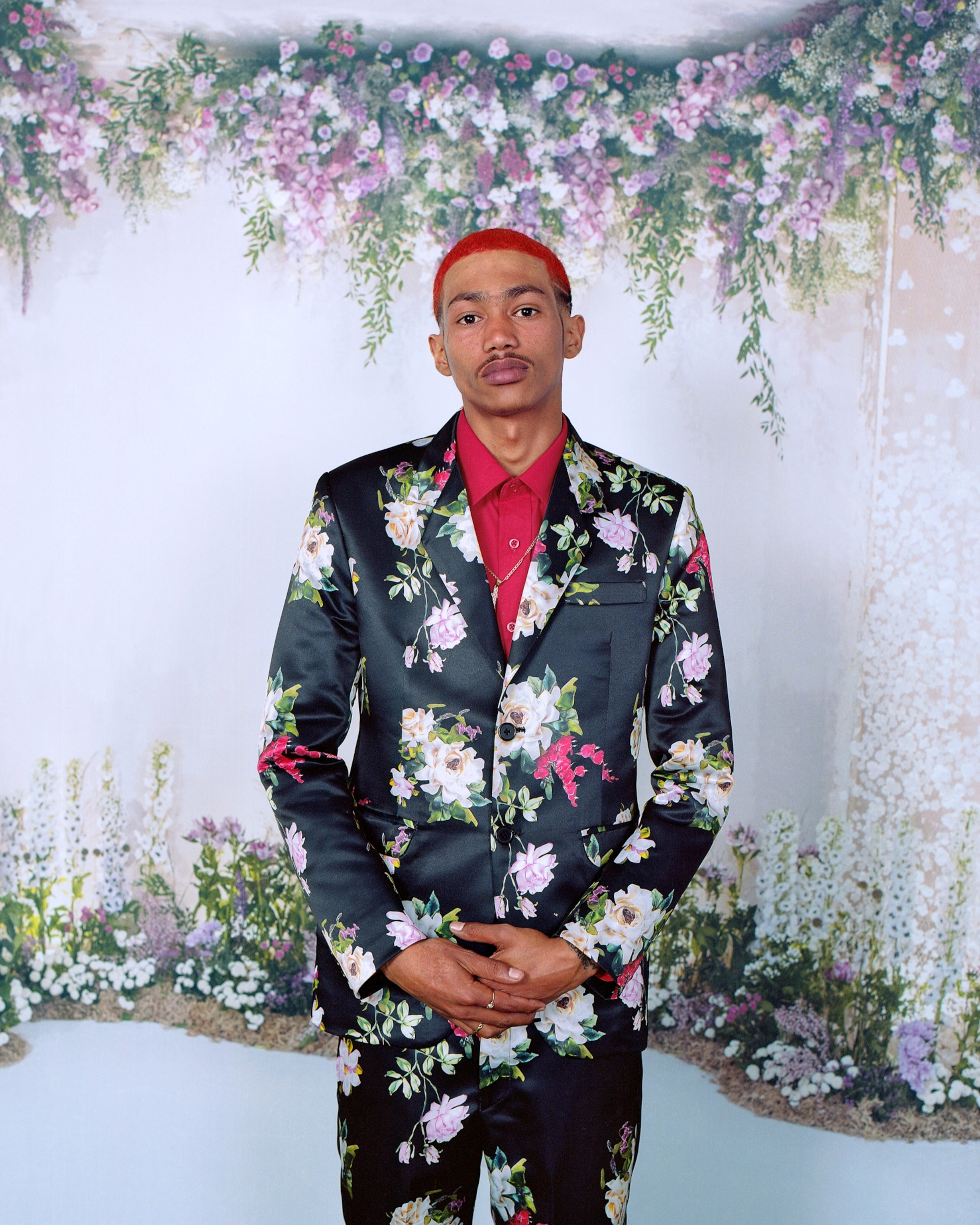 A Black man with red hair wearing a red button down shirt with a floral blazer over it.