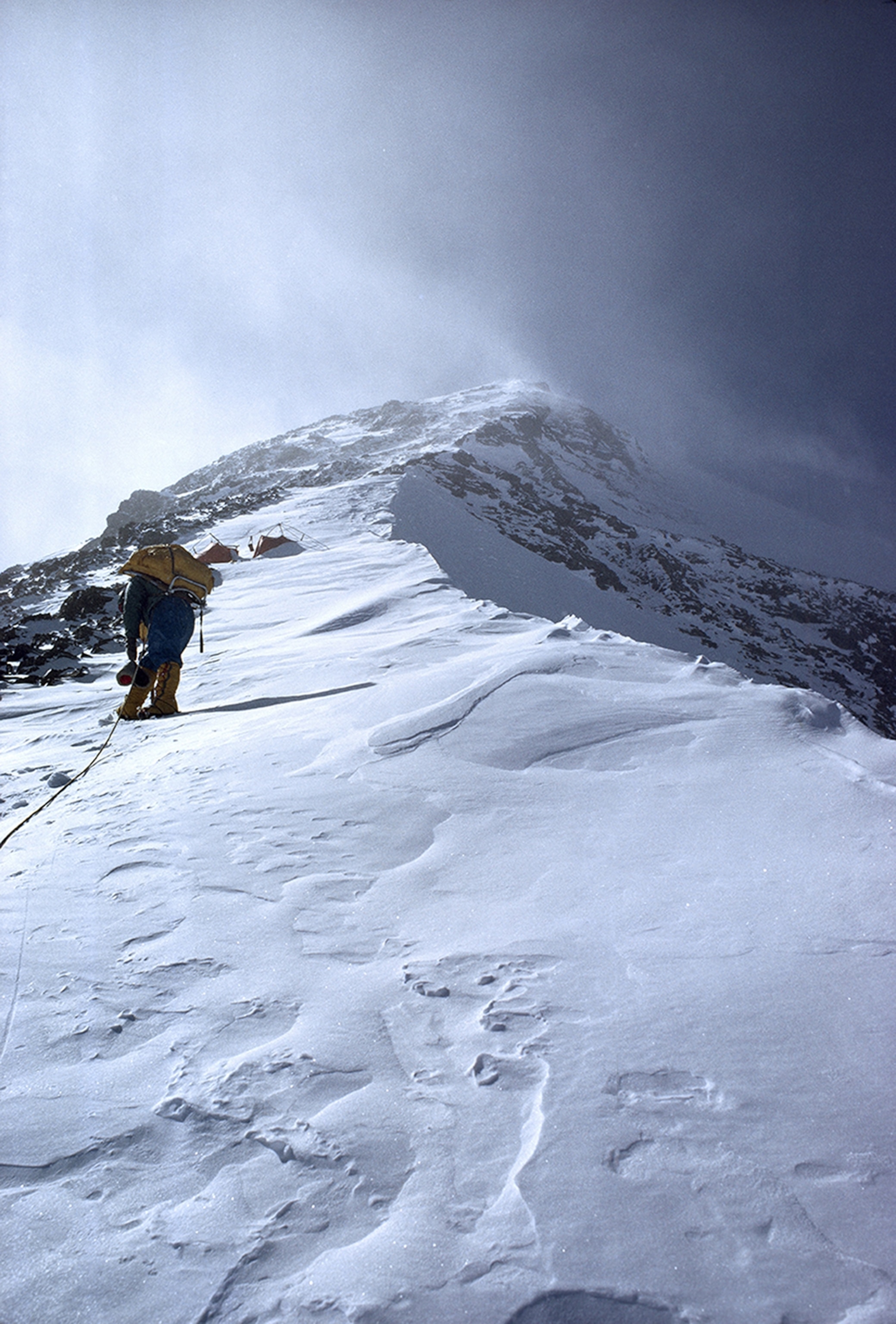 A day from the summit, Jerstad arrives at Camp VI. After taking each short step, he halts for several deep breaths. His rope leads back to Bishop, who took the photograph. Camp consists of the two little Draw-Tite tents half-buried in snow.