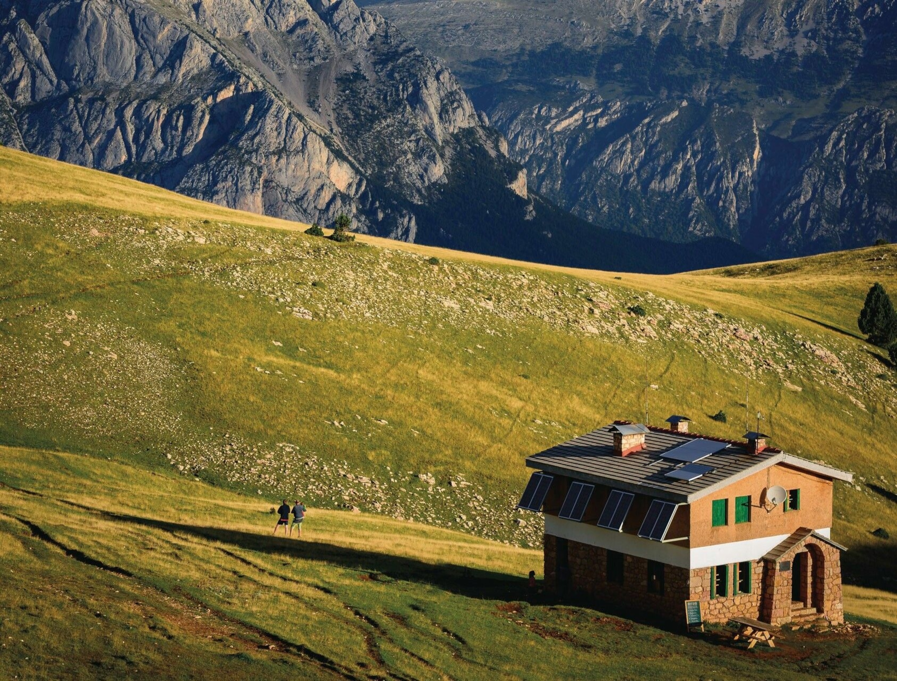 A remote cabin, surrounded by mountains. It is an interesting architectural mix of old stone and modern technology, lined with large solar panels.