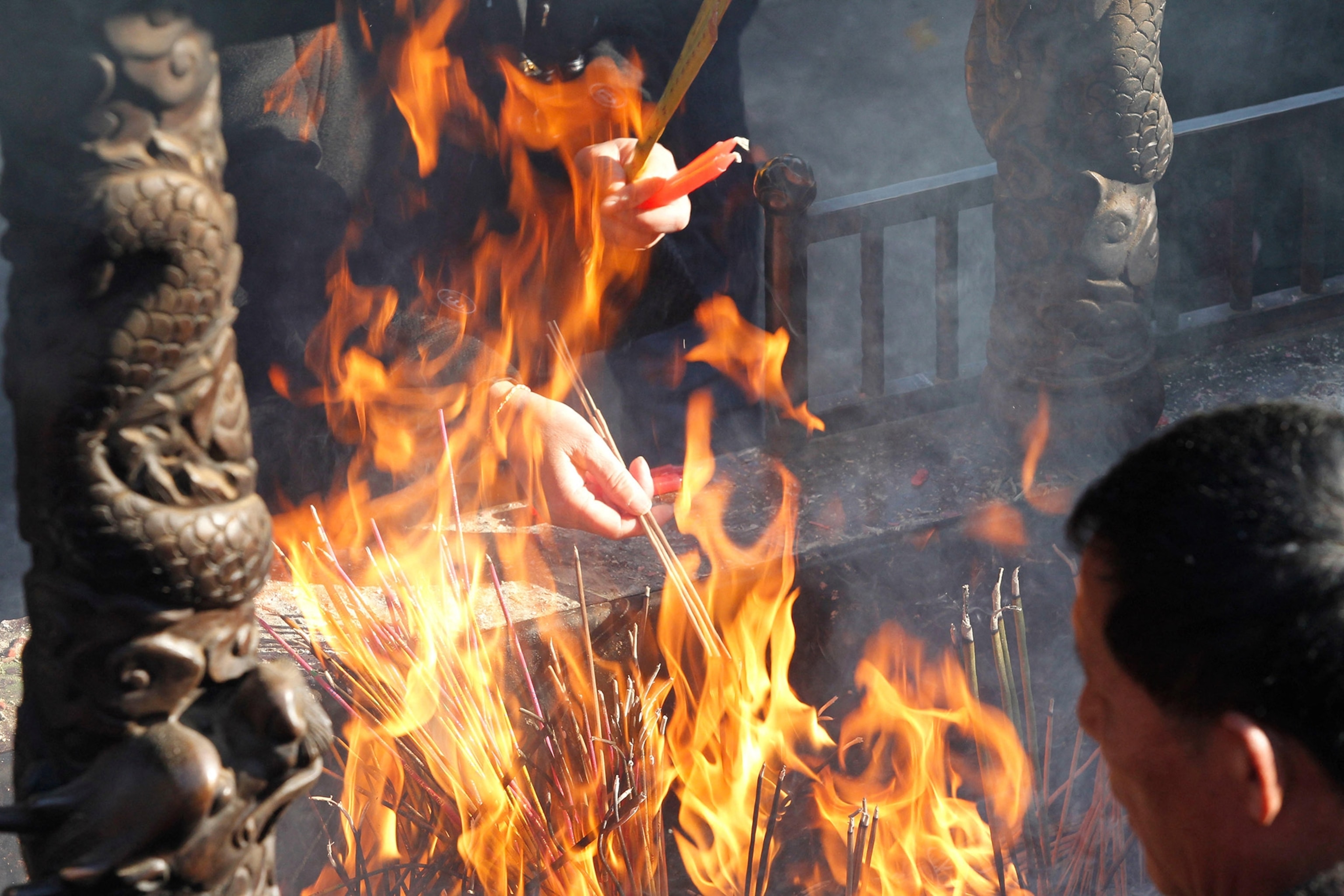 A close view of flames with sticks of incense, multiple hands reach out to light the sticks. A man's head faces away into the fire in the bottom right corner of the frame.