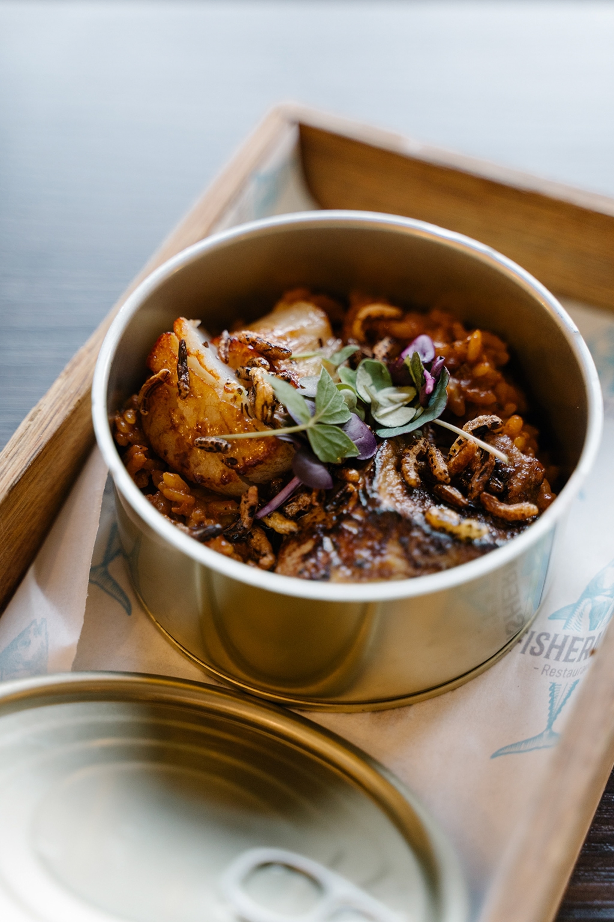 A tin, otherwise used for brined fish, served on a wooden board at a restaurant and containing a fish stew with puffed rice.