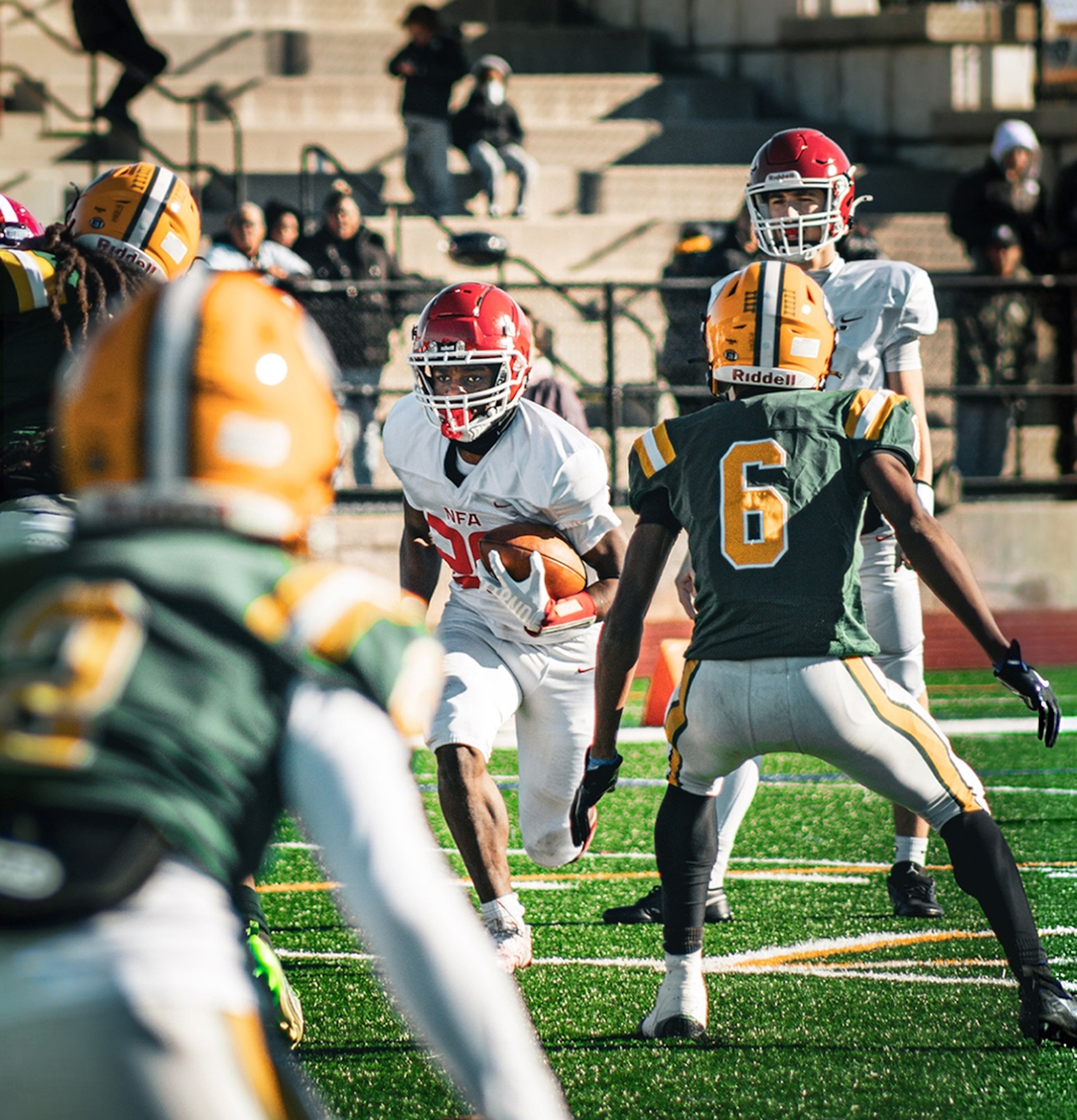 A boy in a white uniform is holding the football and running towards the cameras as two boys in green uniforms stand in a wide-stance facing the player.