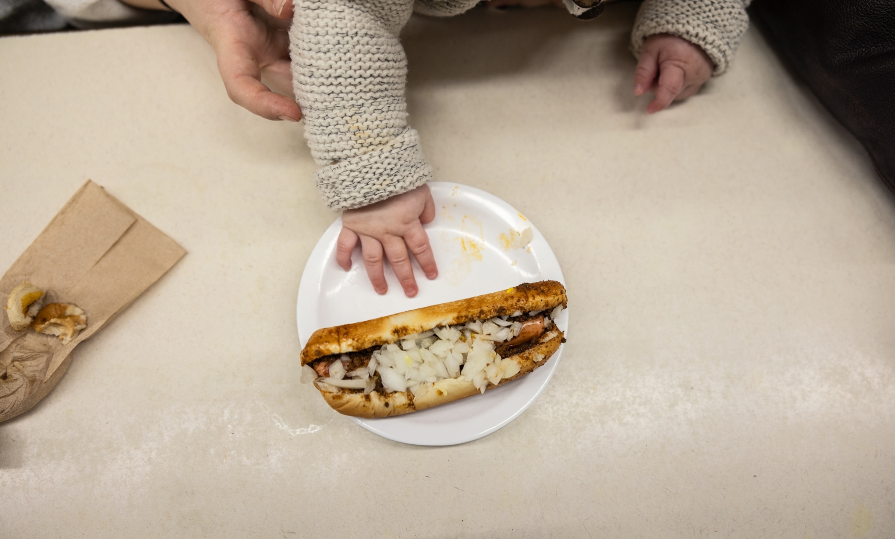 A baby's hand reaches for a hot dog on a white plate.