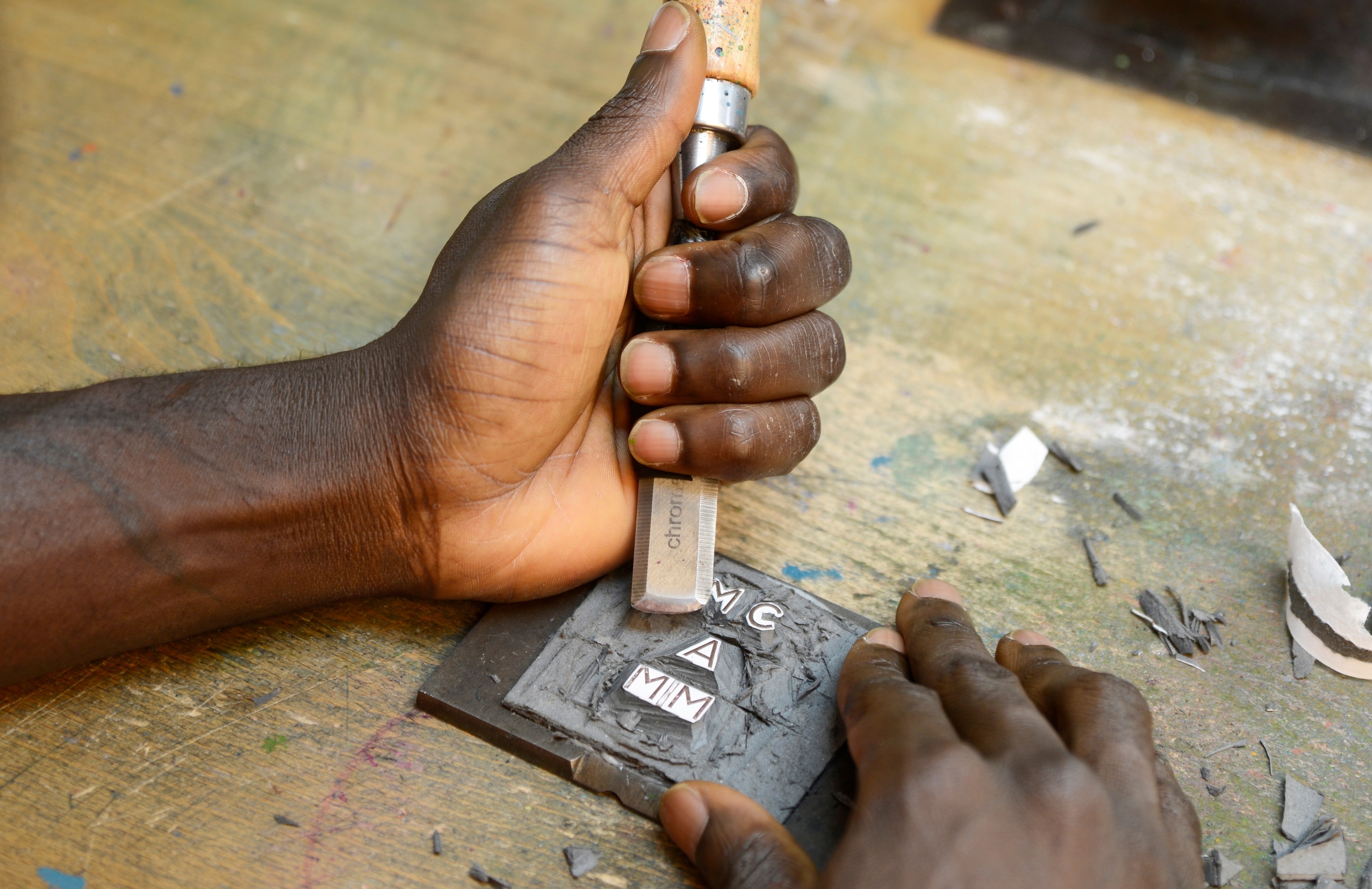 Two hands hold a chisel and a small plate with letters on it as the creator chisels away the material around the letters.