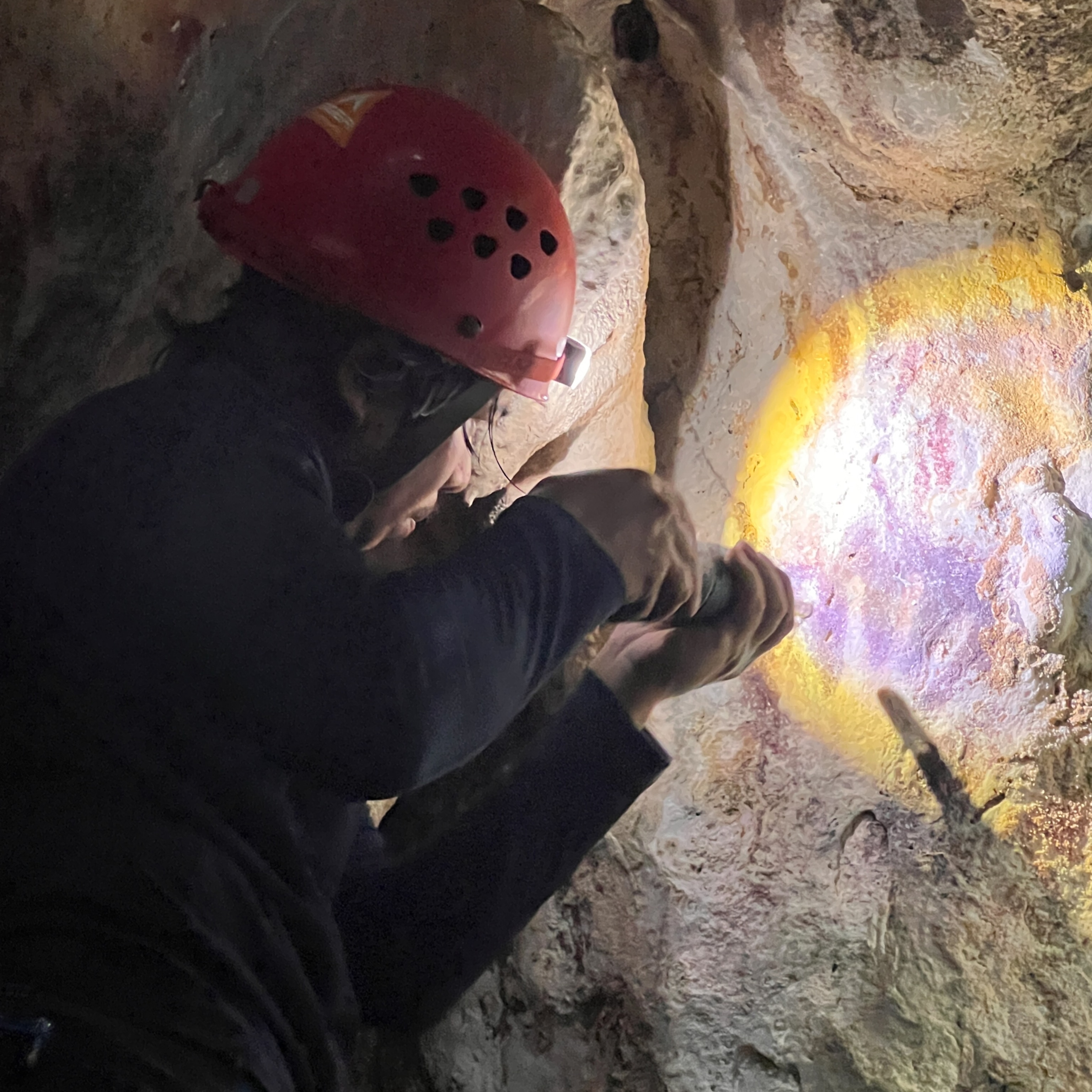 A person wearing a red helmet examines a cave wall closely, using a special flashlight to reveal the faint remains of hand stencil cave art