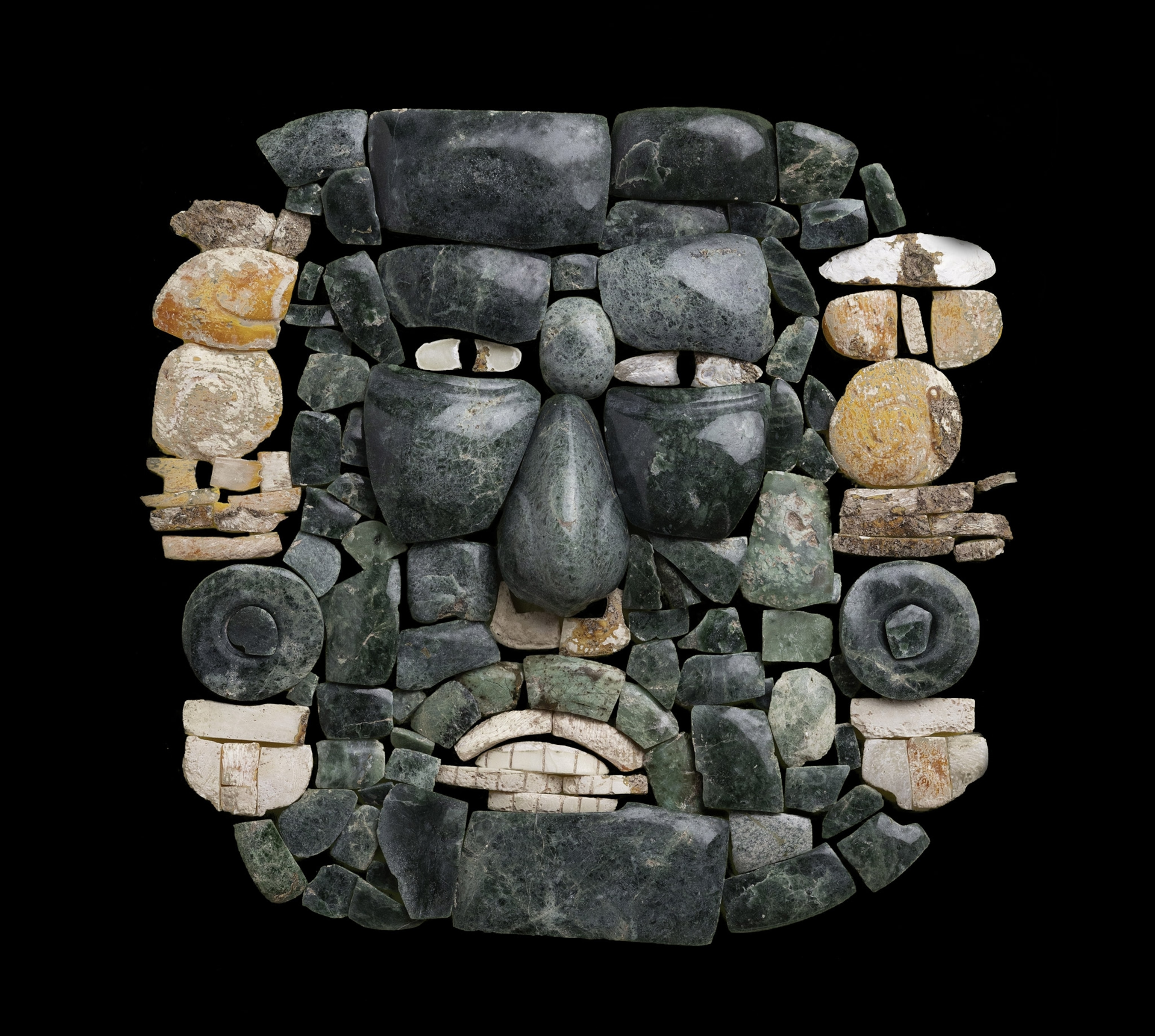 Dozens of pieces of green jade and white shell assembled into a mosaic resembling a face, against a black background