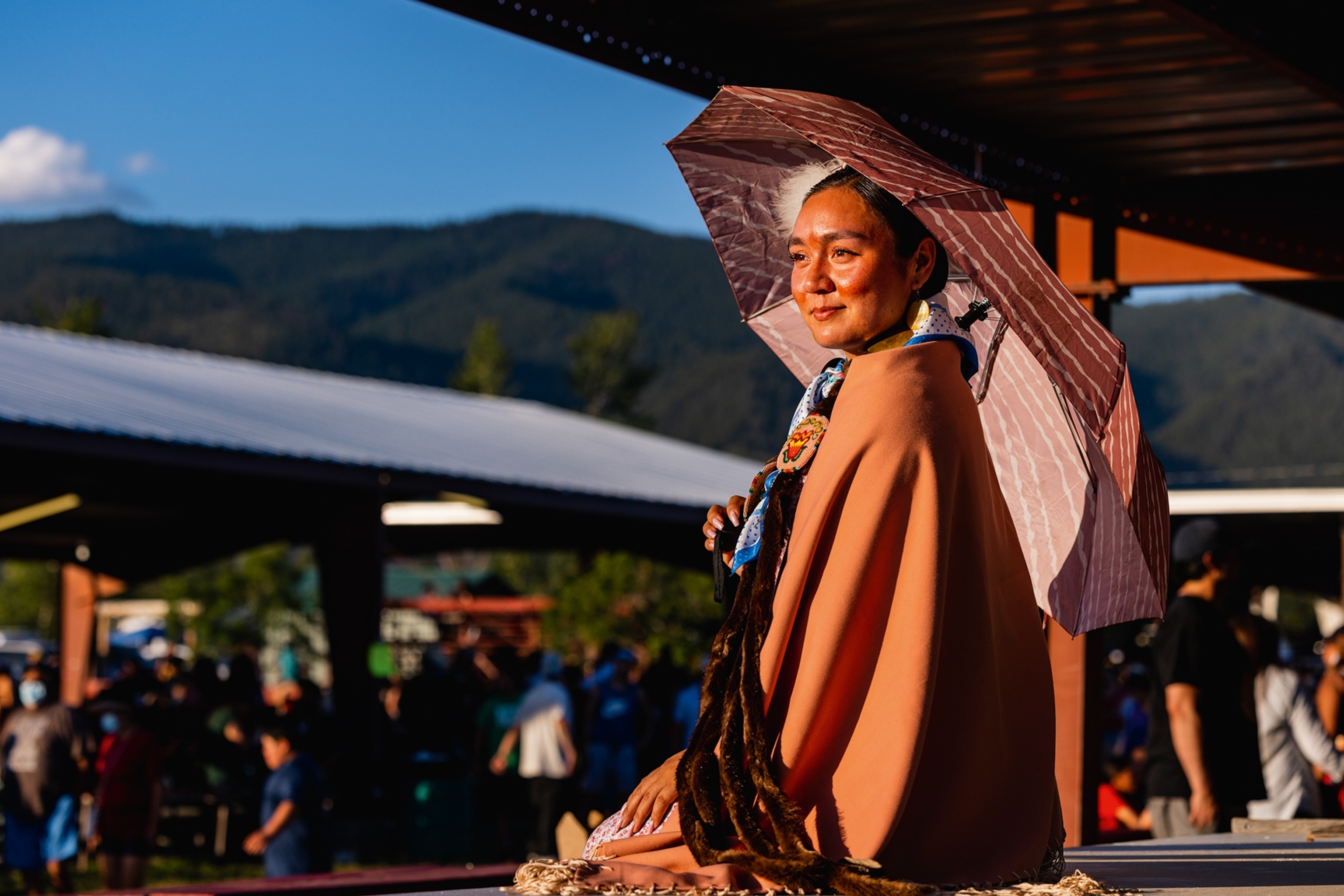 A Native American woman sitting on a wall overlooking the festivities, the sun illuminating her flushed skin as she holds an umbrella behind her.