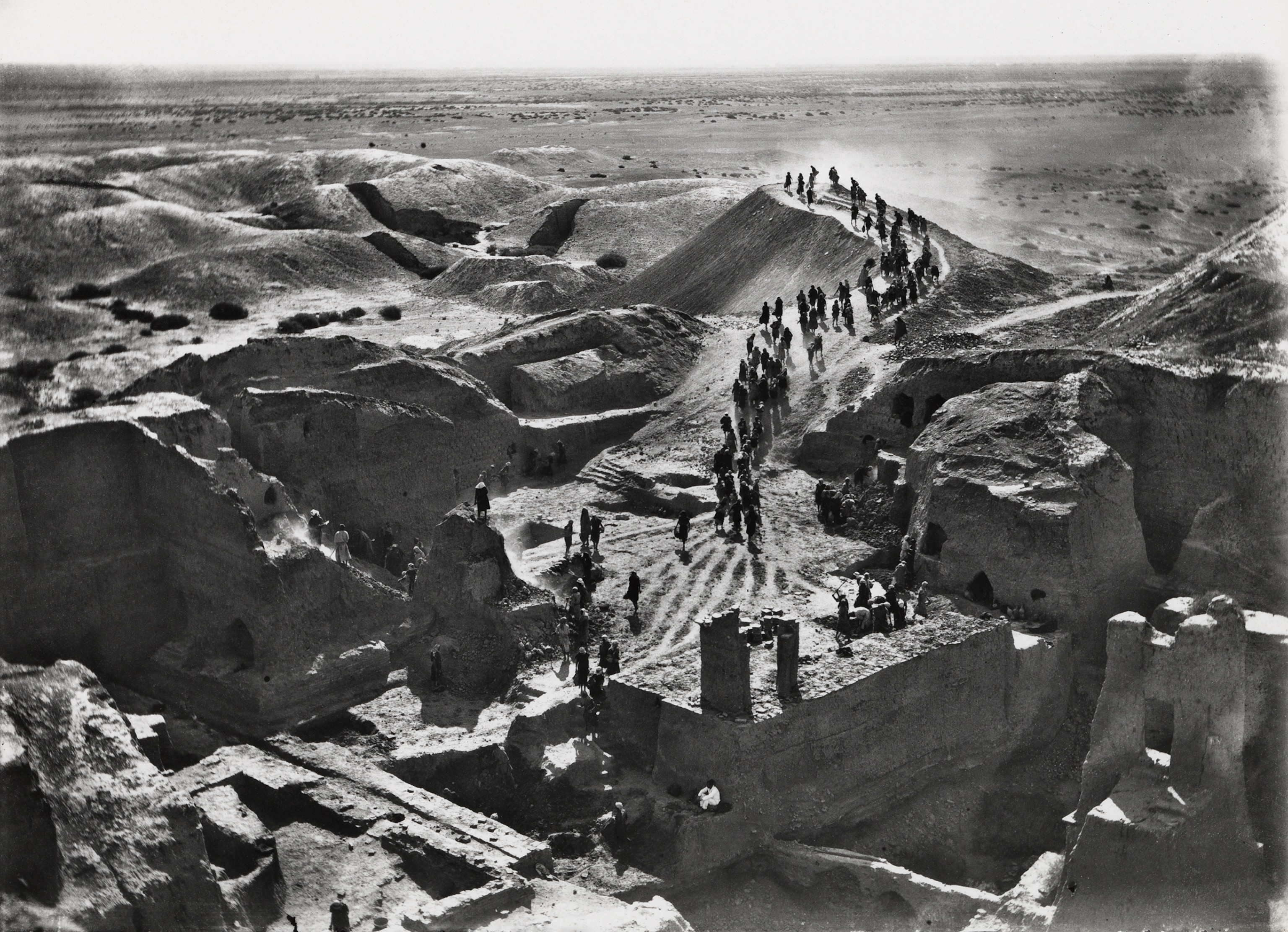 Black and white photograph of the excavations of Temple enclosure as seen from top of Ziggurat, with people walking down a winding path, Nippur, Iraq.