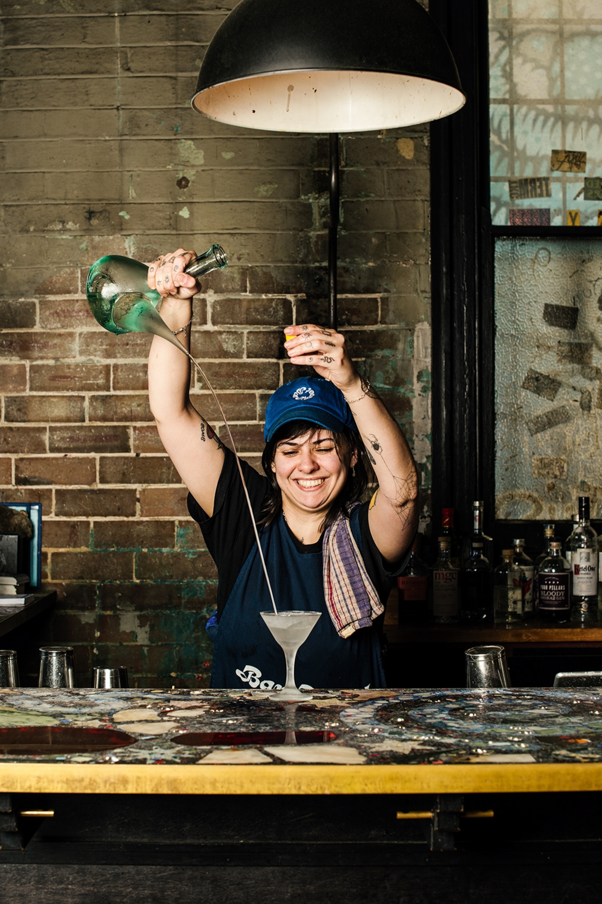 A young female bartender pouring a martini from a glass carafe with stretched out arms while smiling.
