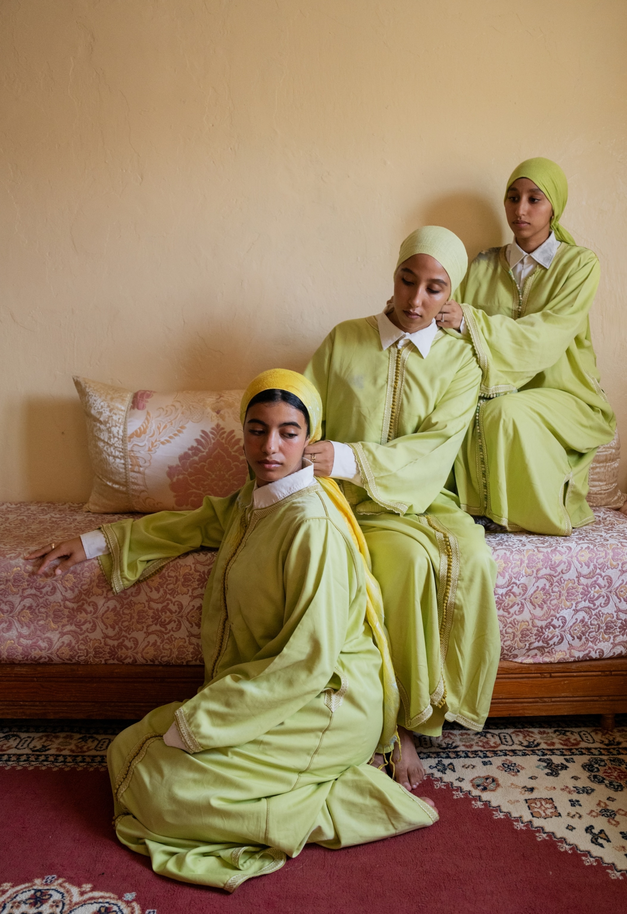 A woman sits on the ground and two sit on the couch, each facing the back of the person next to them and actively fastening their scarf.