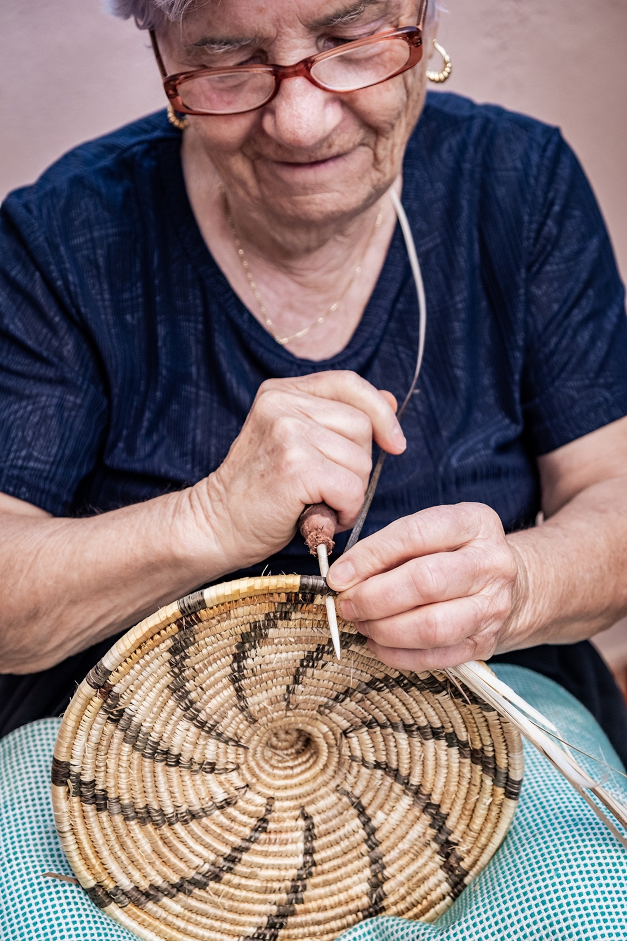 An elderly woman holding weaving tools and a basket in progress.