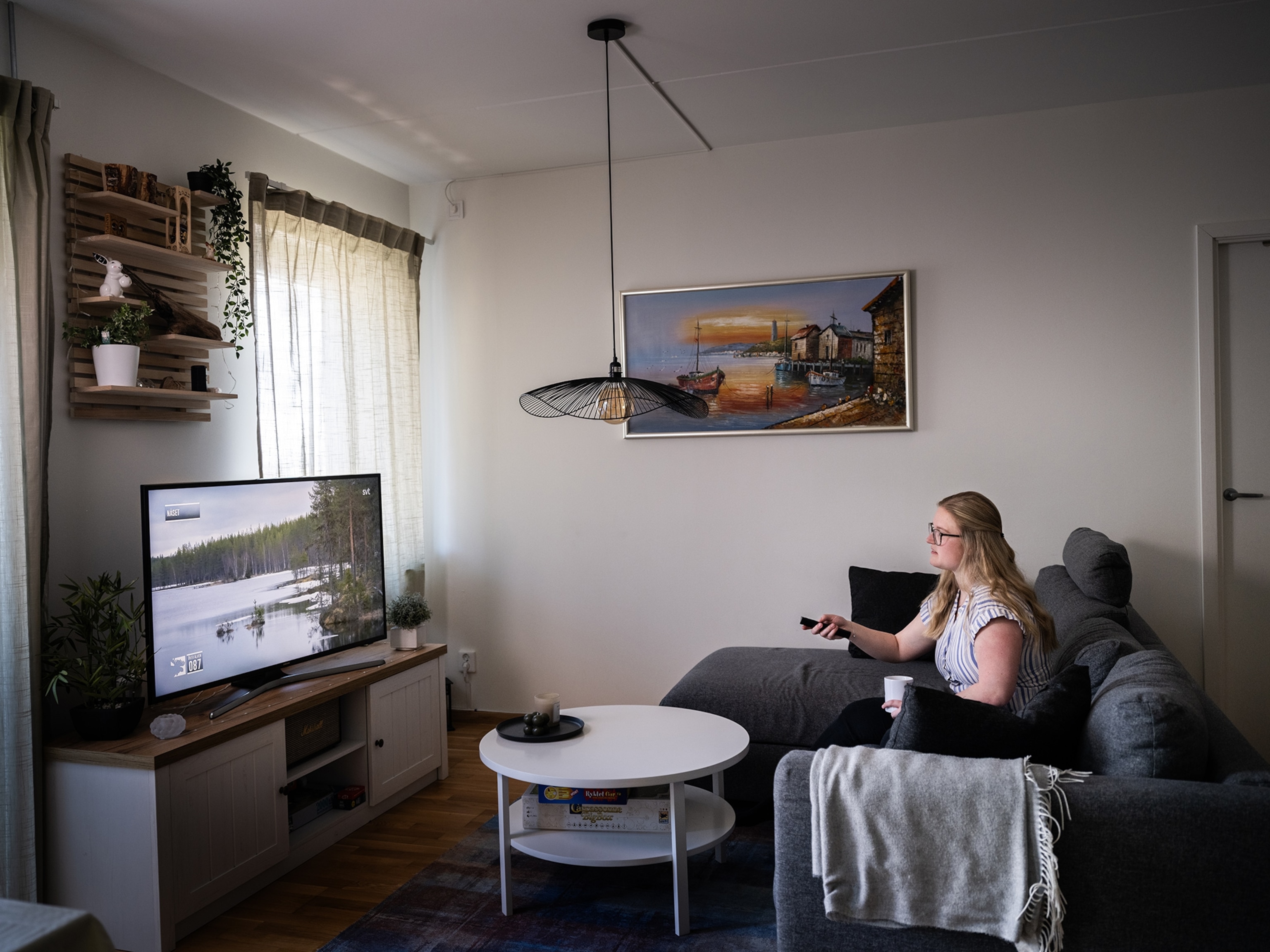 a blond woman in a white shirt sits on a grey couch in her living room aiming the TV remote at the TV. The TV is showing a nature scene of a riverbank and trees