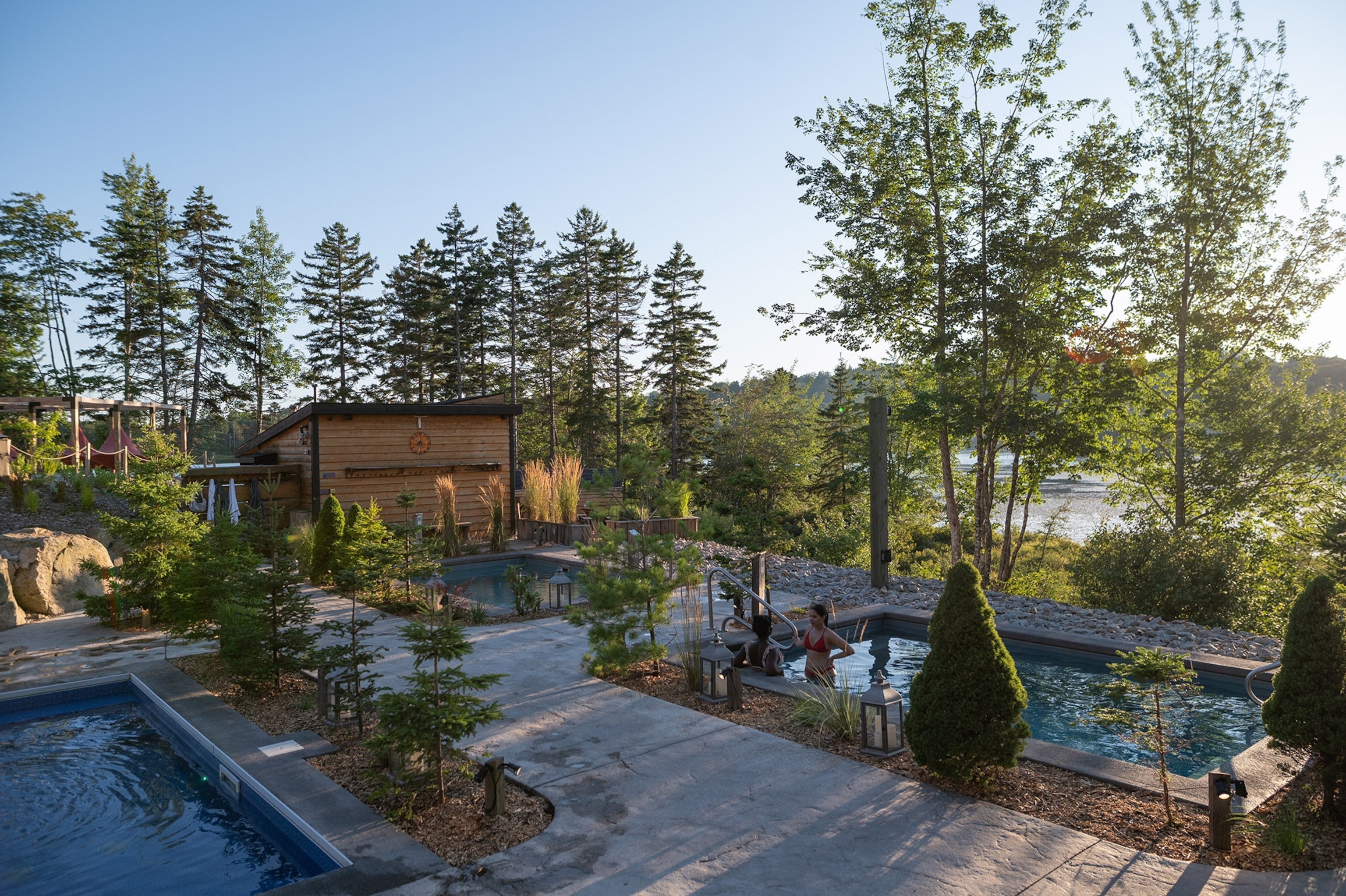Outdoor pool deck with a small wooden shed, two small pools, surrounded by trees. In one pool, two women stand partially submerged