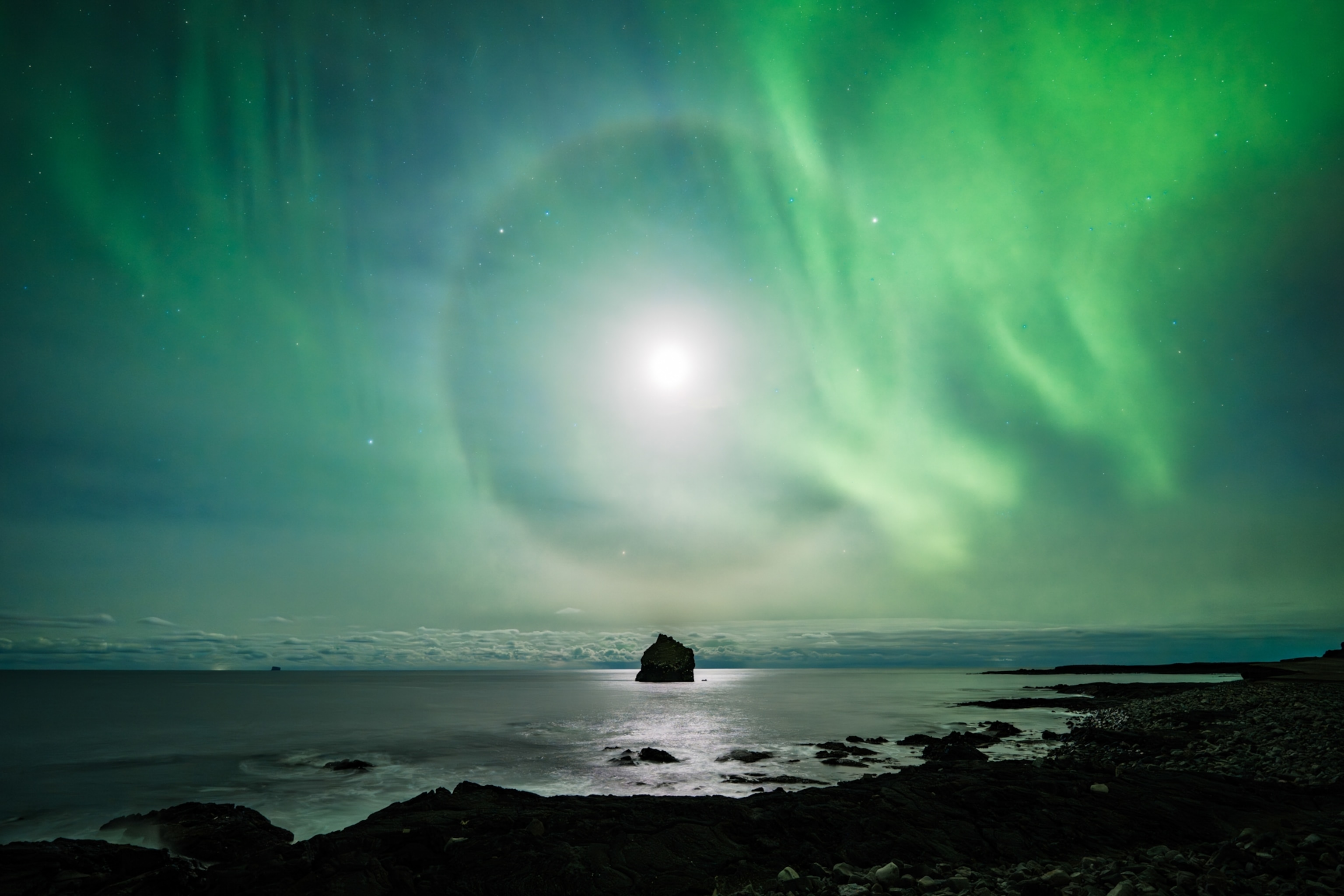 A serene coastal scene under the northern lights. A glowing halo surrounds the moon, casting a green aurora over the sea's rocky shore