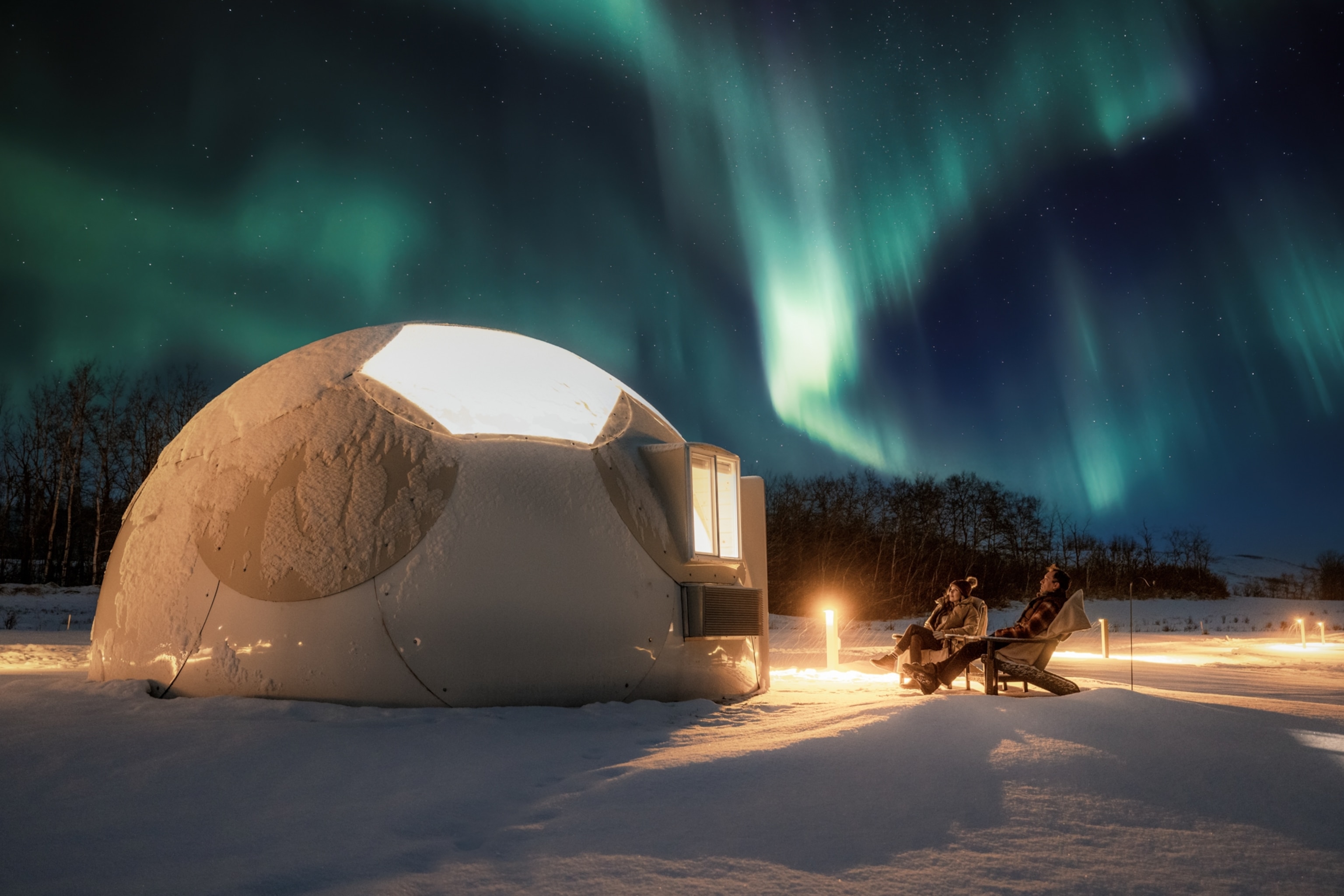 A couple seated outside a dome cabin, beneath the Northern Lights in the night sky