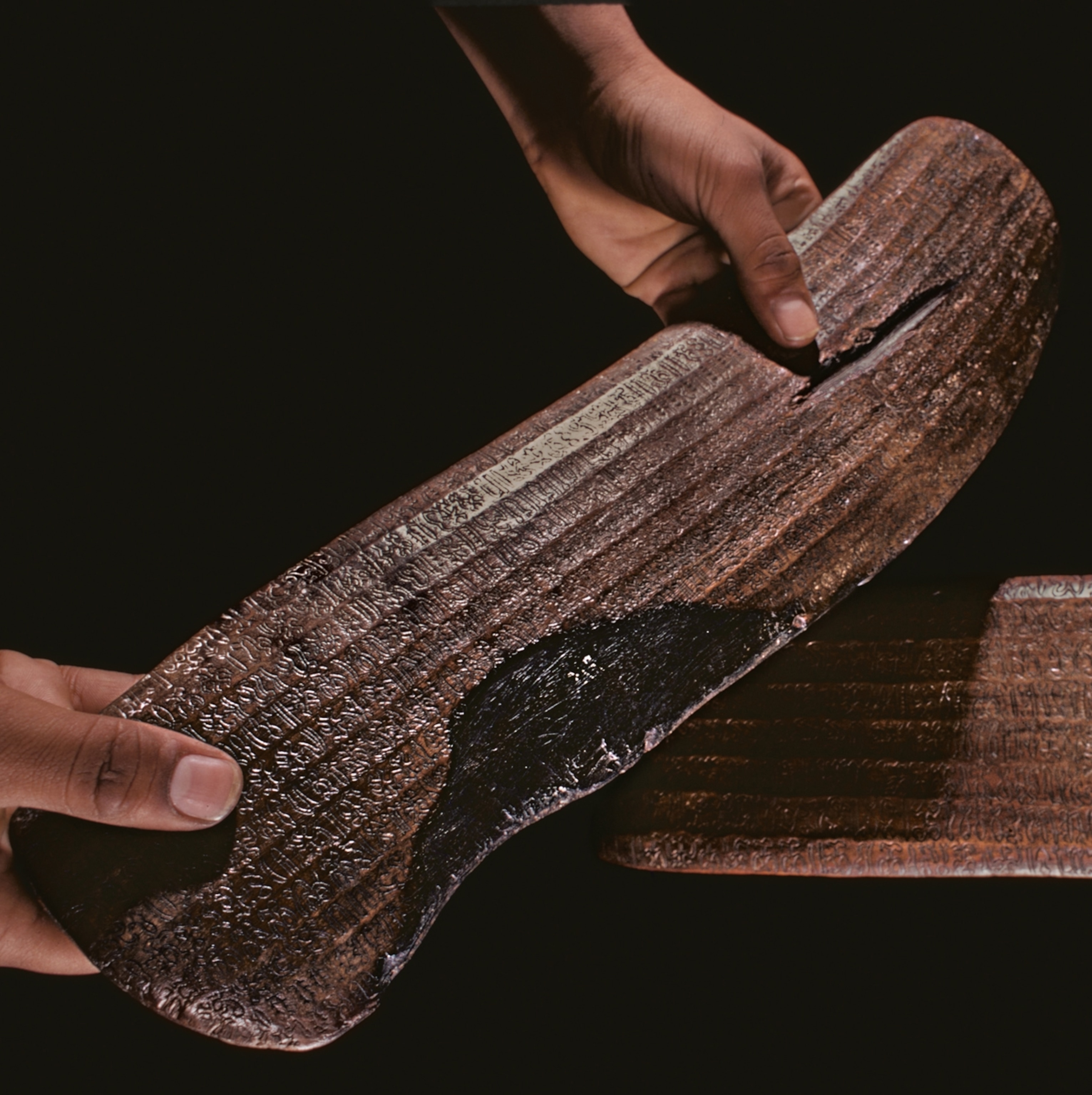 Two hands hold a long wooden object with writing carved into it.