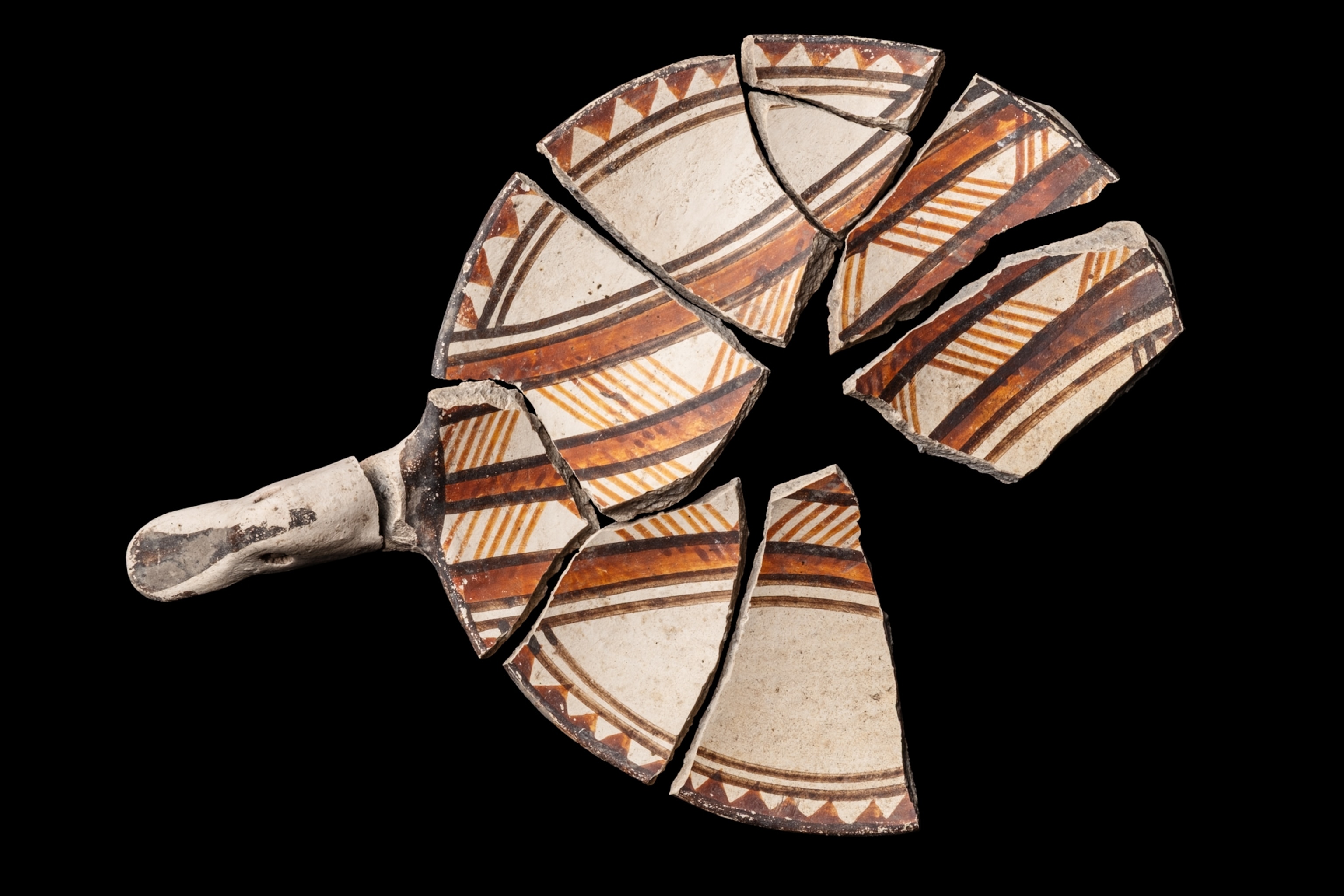Fragmented bowl with a converging rim, concave body, flat base, and a handle featuring a modeled ornithomorphic motif. The vessel is decorated with painted geometric designs, including triangular figures hanging from the rim, as well as parallel lines, bands, and zigzag patterns.