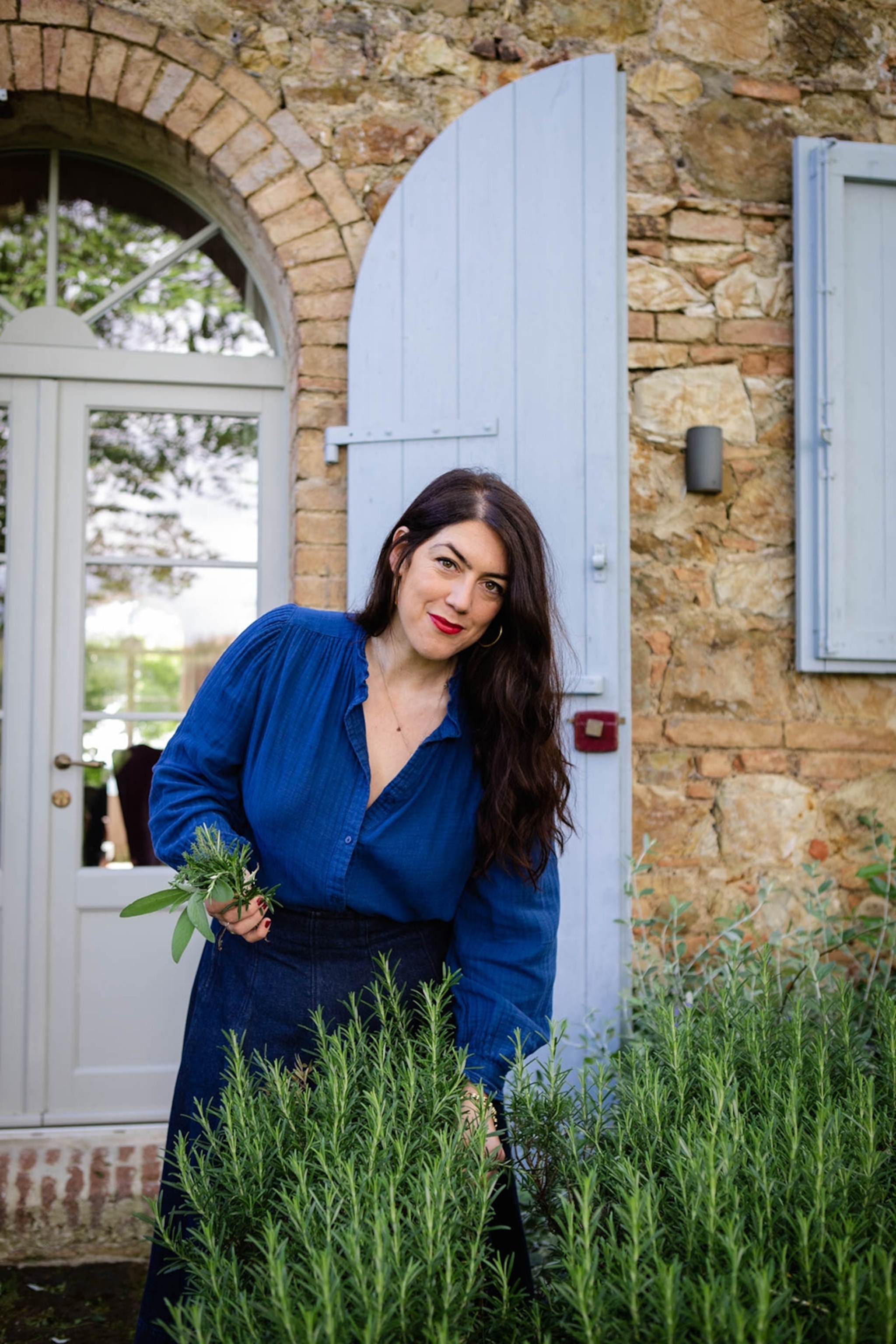 A casual portrait of a long-haired, smiling woman outside of an Italian house picking rosemary from a bush.