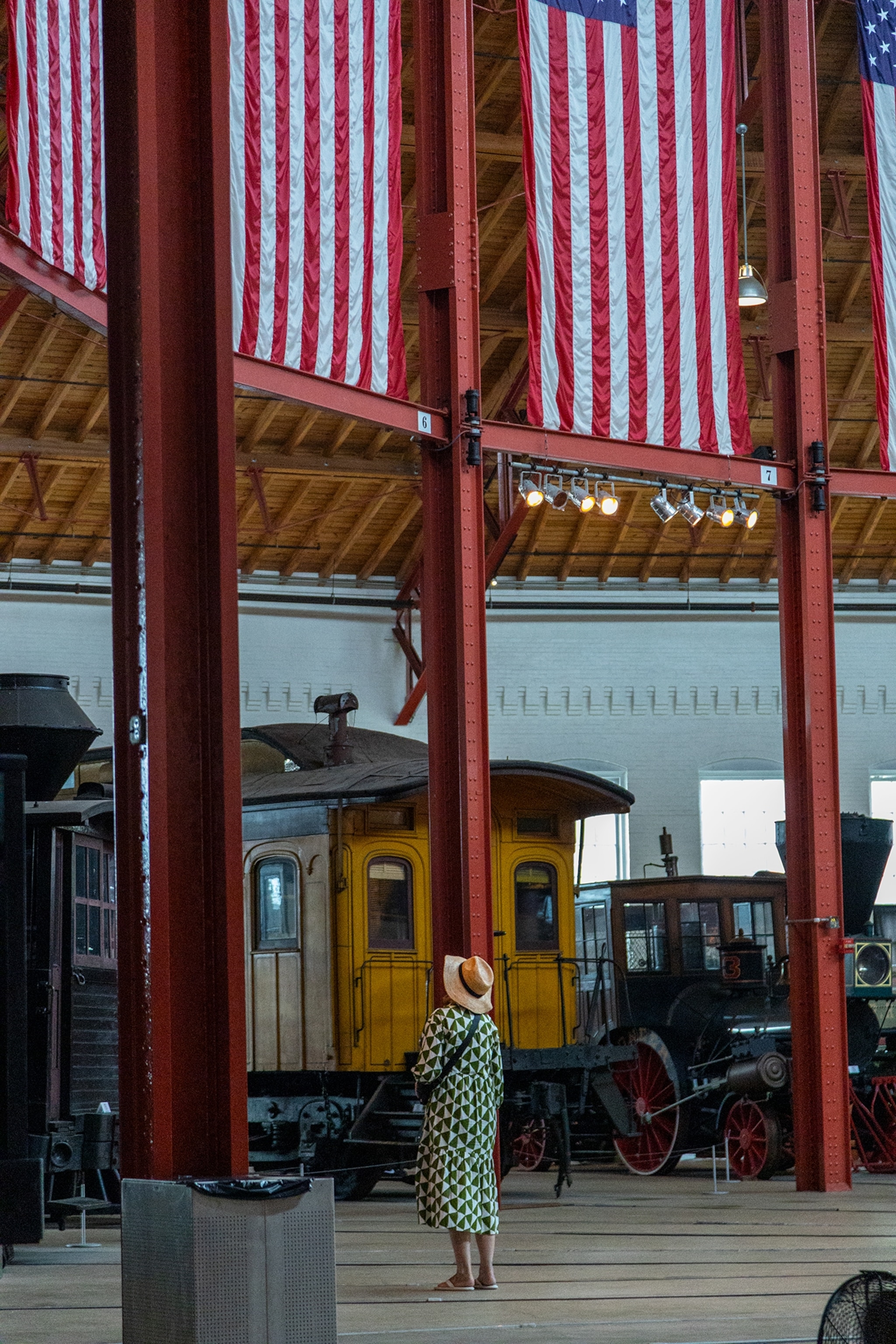 A person wearing a patterned outfit and hat stands inside a historic train museum, looking at vintage railcars and locomotives displayed beneath large American flags hanging from the ceiling.