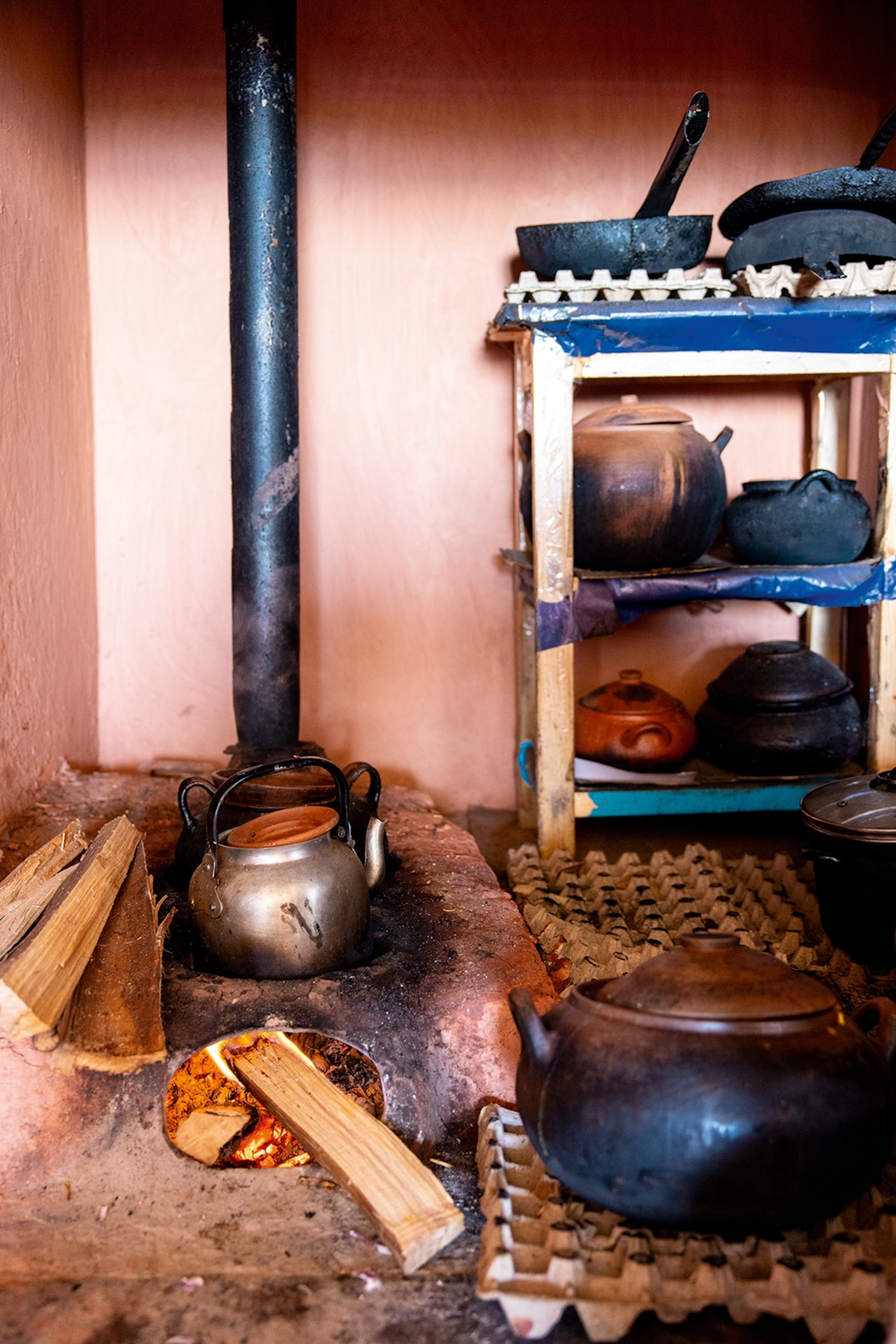 The interiors of a simple farm kitchen with a stove and kettle on the floor and a shelving unit to the side.