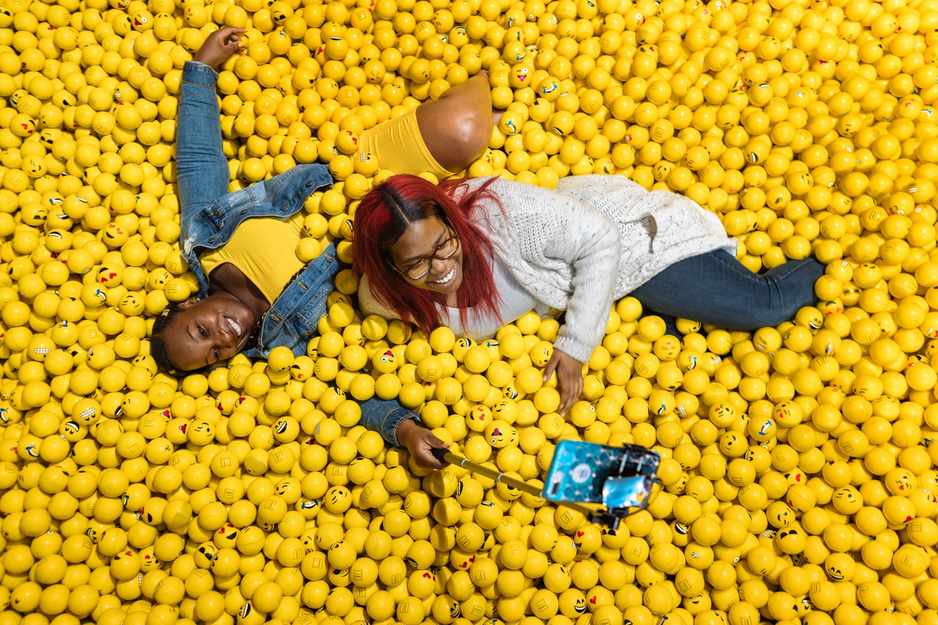 two people lying in a yellow ball pit of emojis, taking a selfie