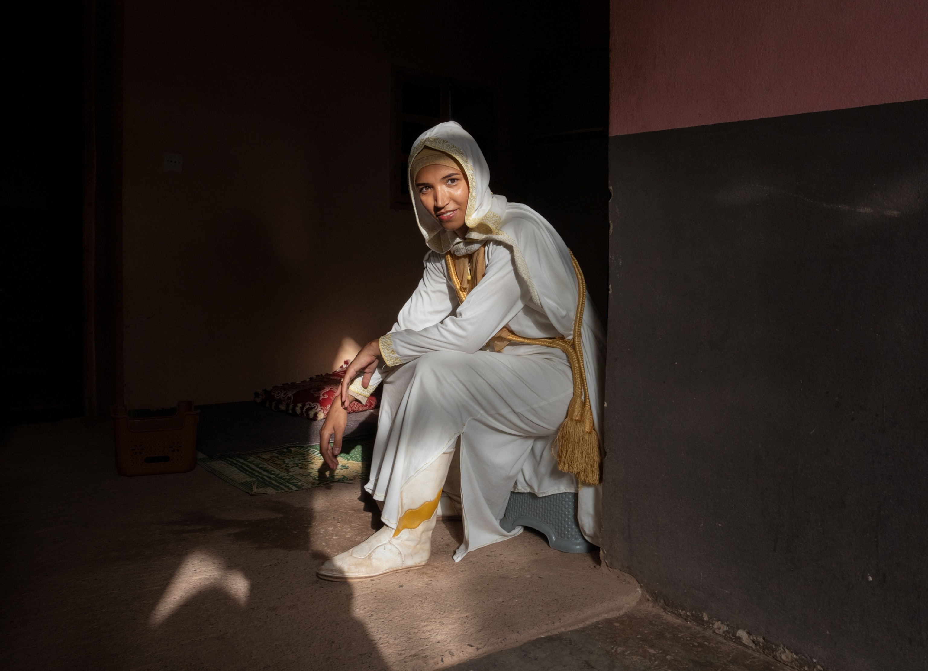 A woman wearing white sits on a step in a dark room as the side illuminates her.