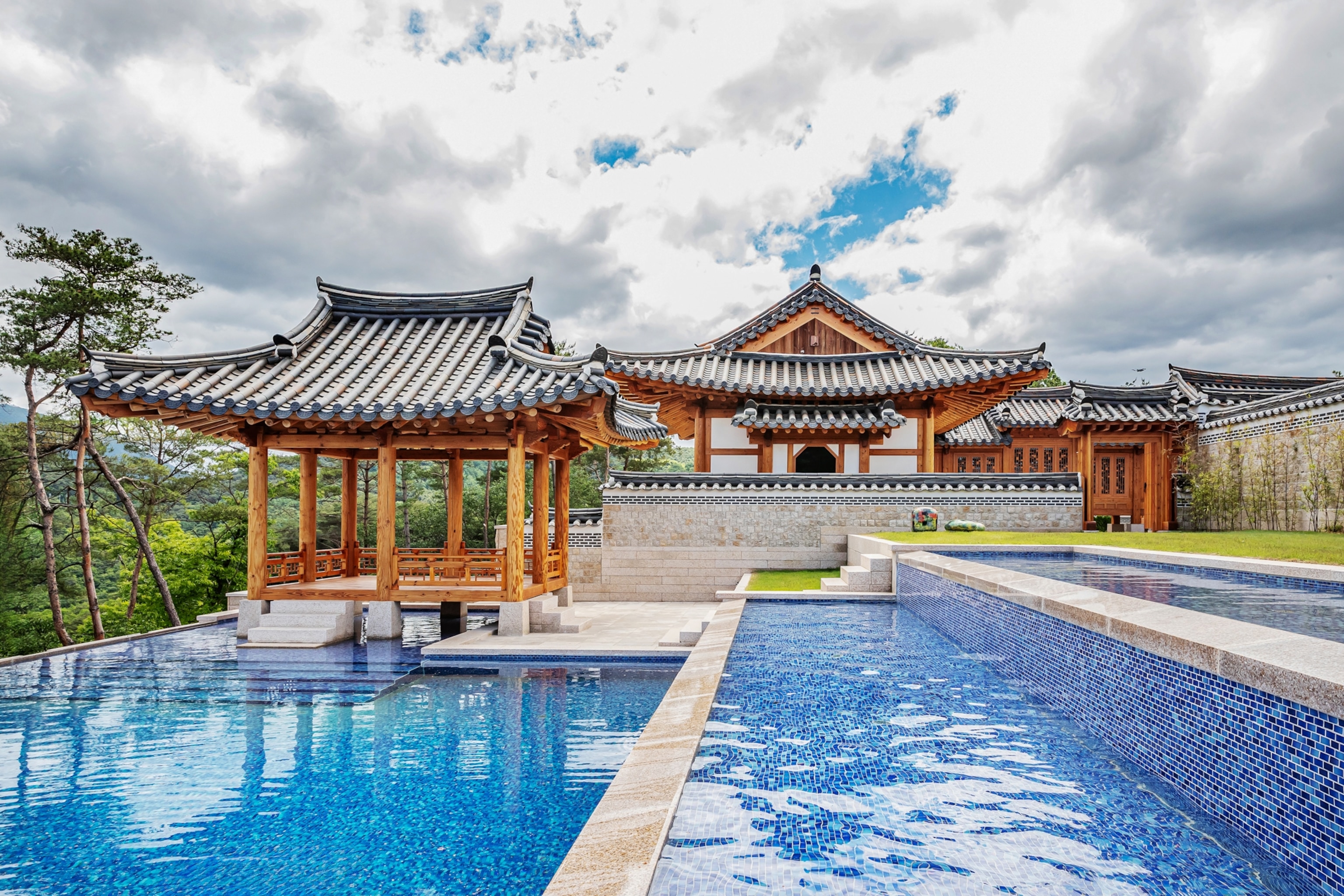 A traditional Korean-style building with tiled roofs and wooden pavilions surrounds a large blue swimming pool under a cloudy sky.