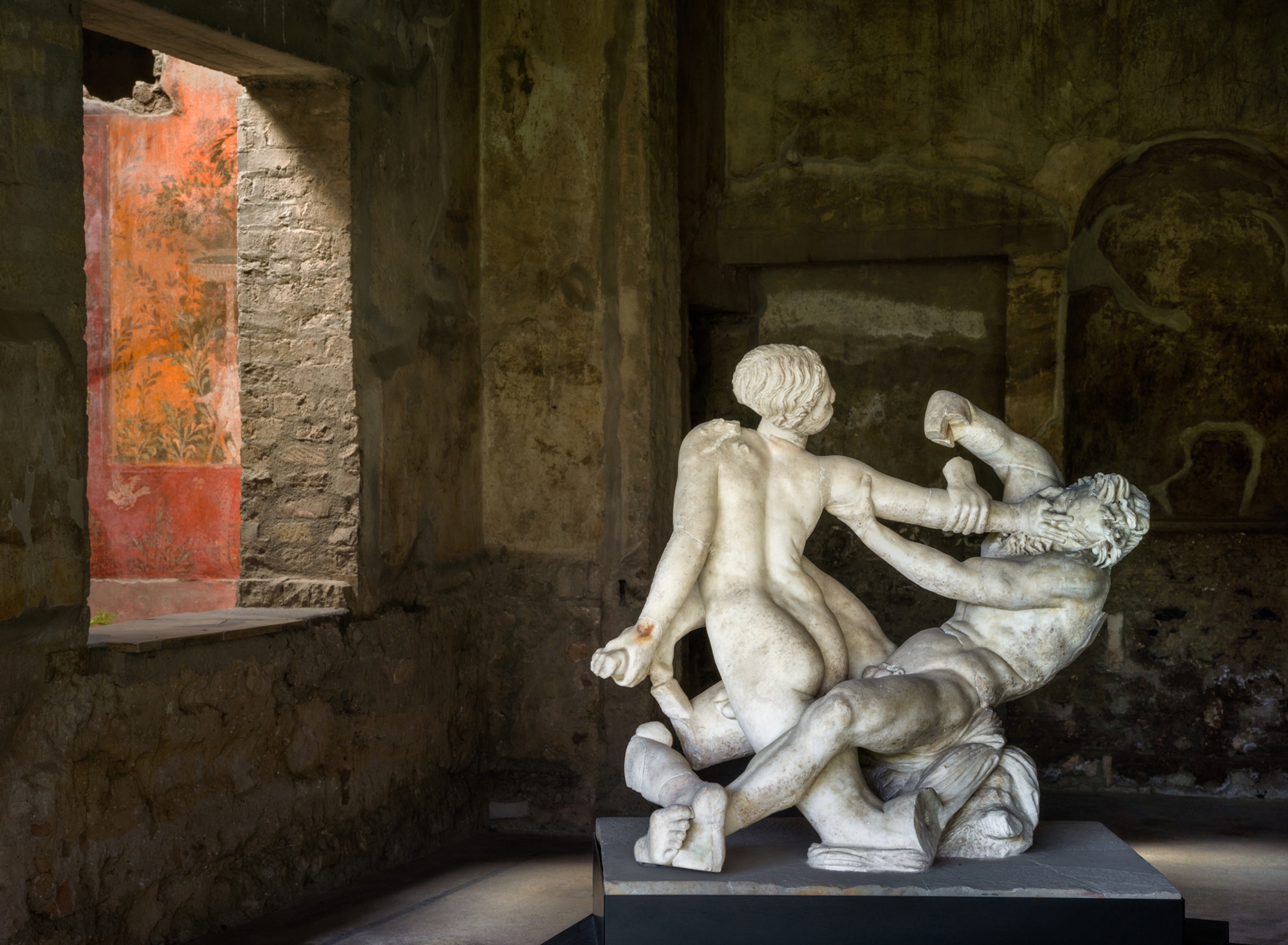 Marble statues that are fighting stand in a room with a window facing it.