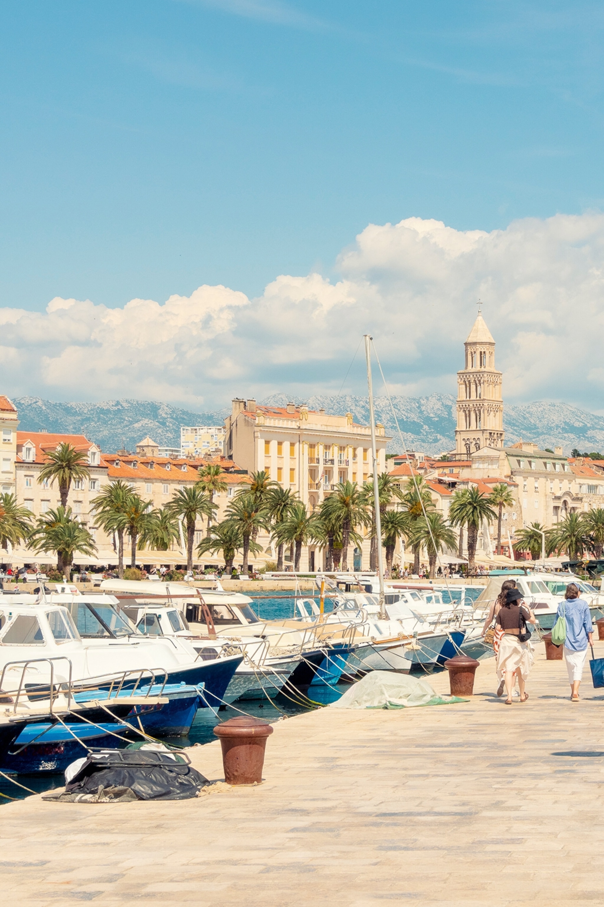 A marina harbour in the sun with boats anchored on either side and a palace tower in the distance.