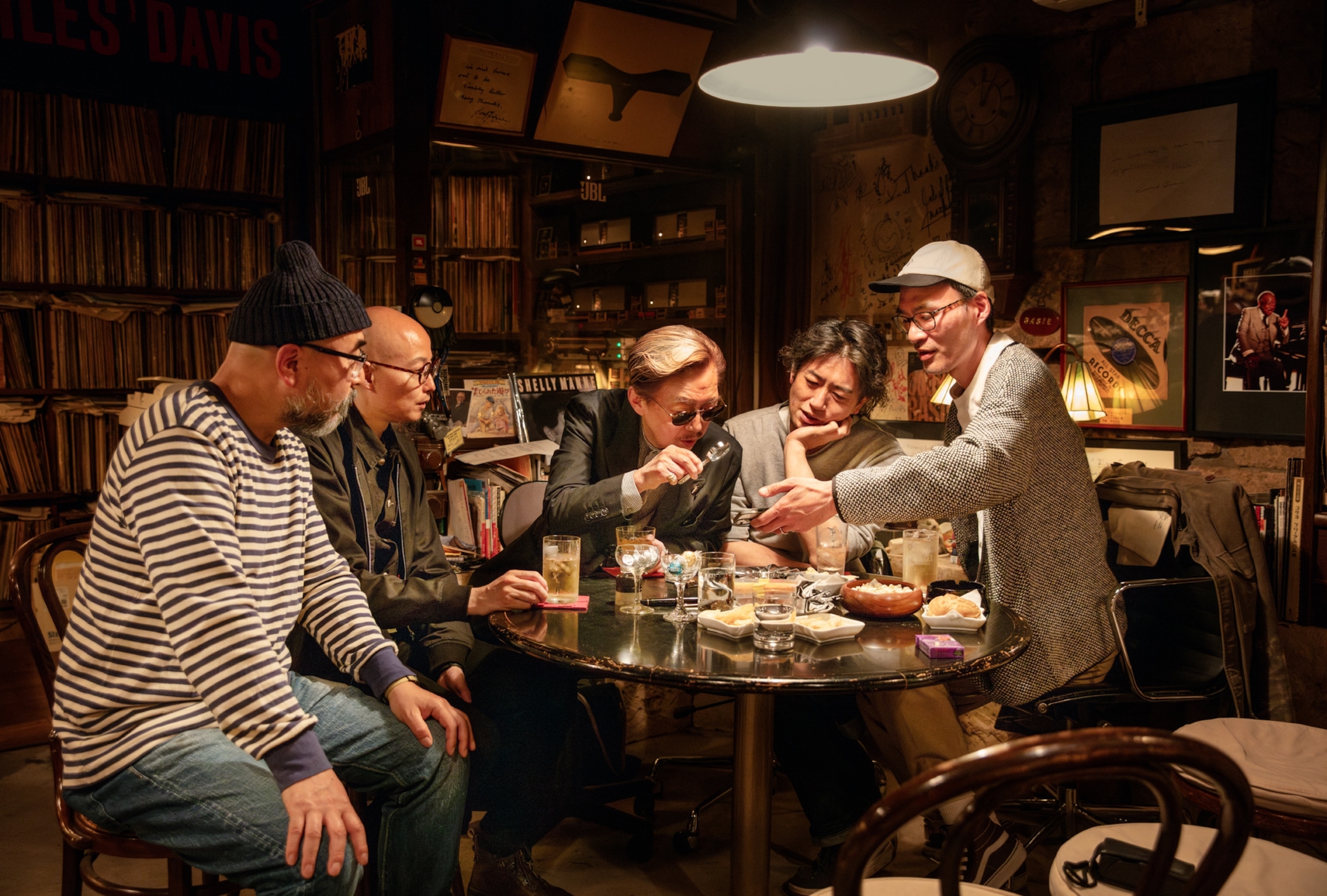 Five Japanese men hover over a round table with drinks and snacks.