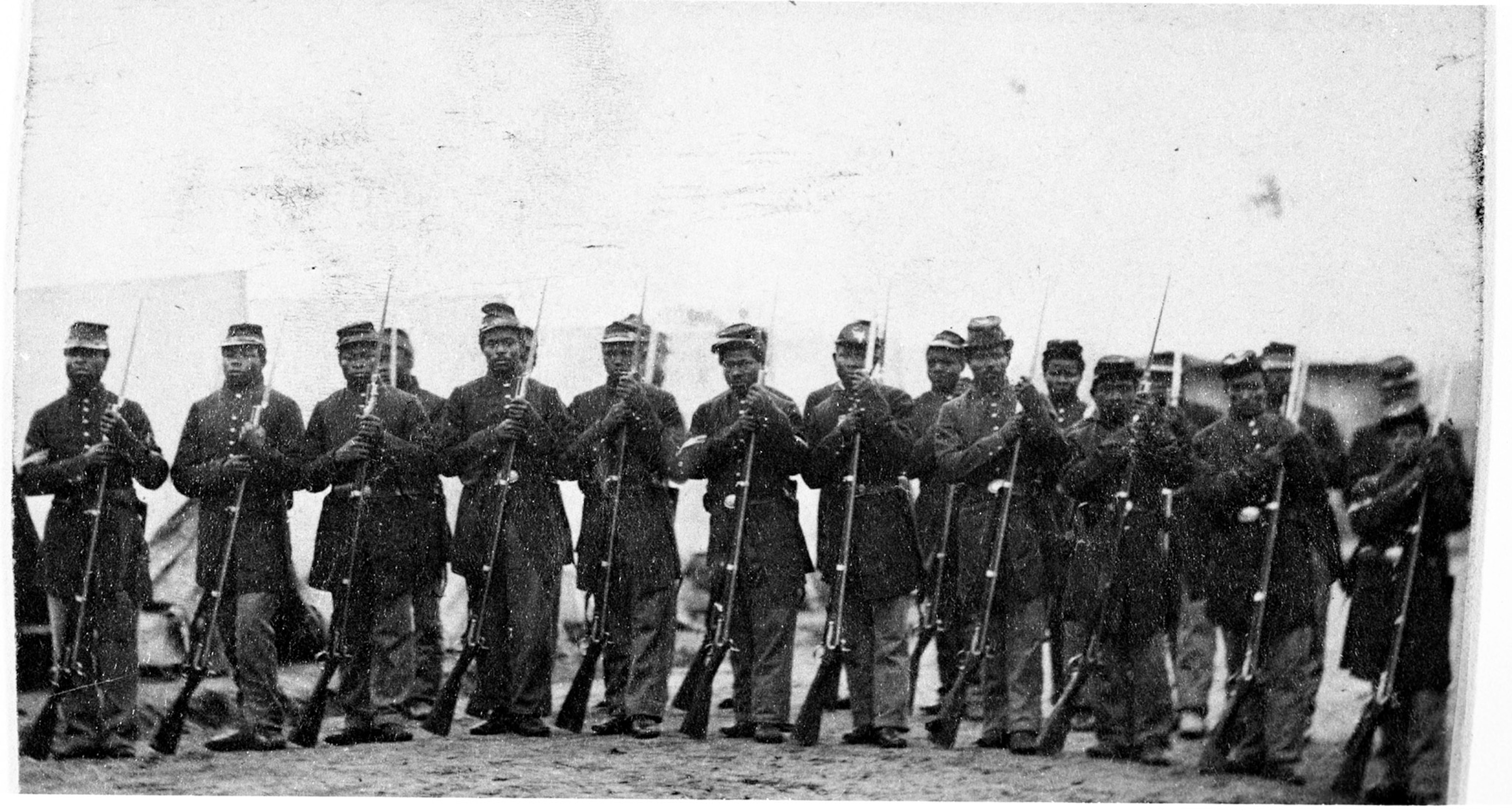 A black and white archival image of African-American military troops standing in line looking at the camera.