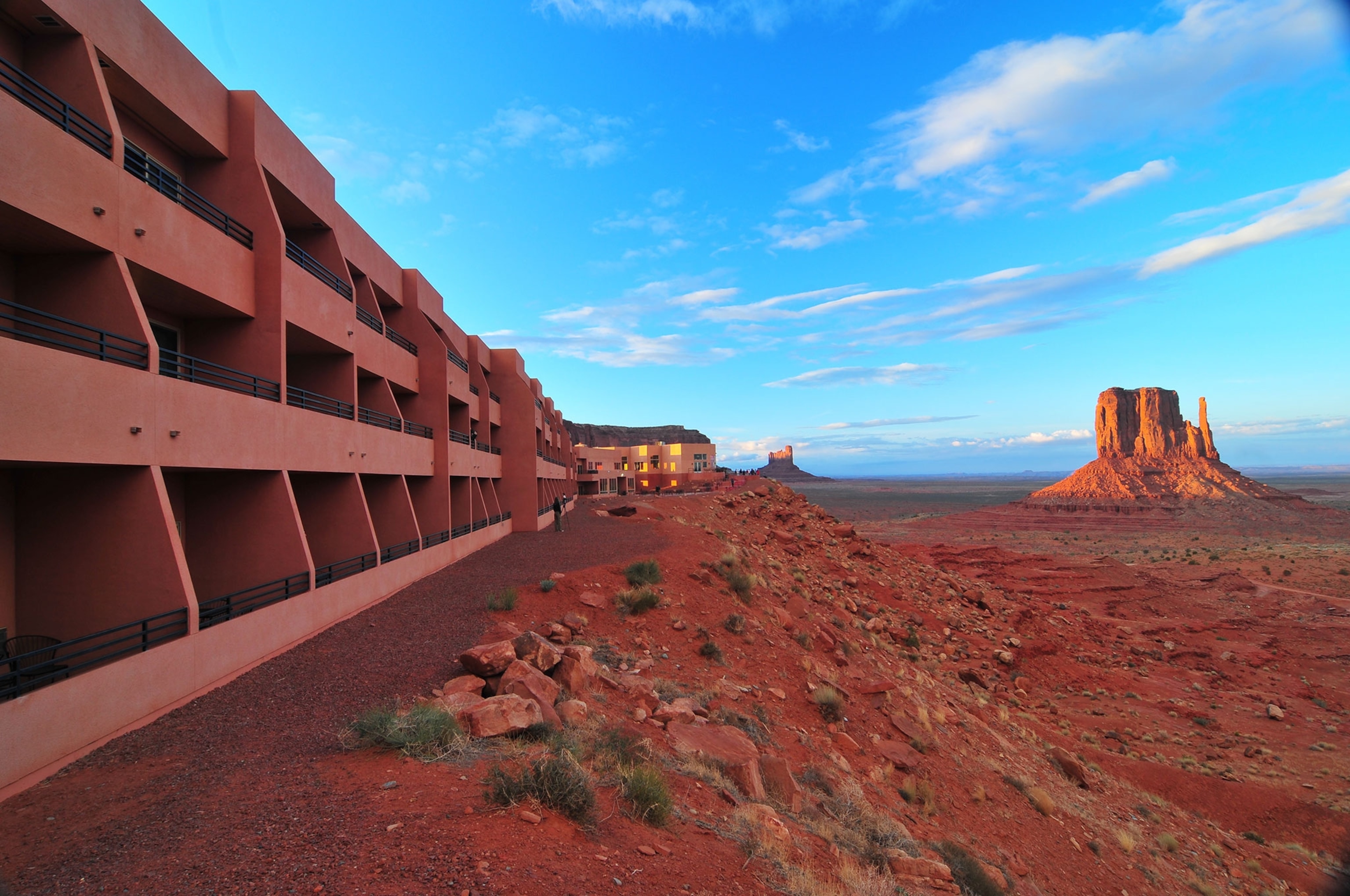 To the left, a redish clay, adobe style building with three floors that extends into the distance on a cliff. In the distance is a desert, with a redish clay rock structure. The sky is blue and the sun is setting.