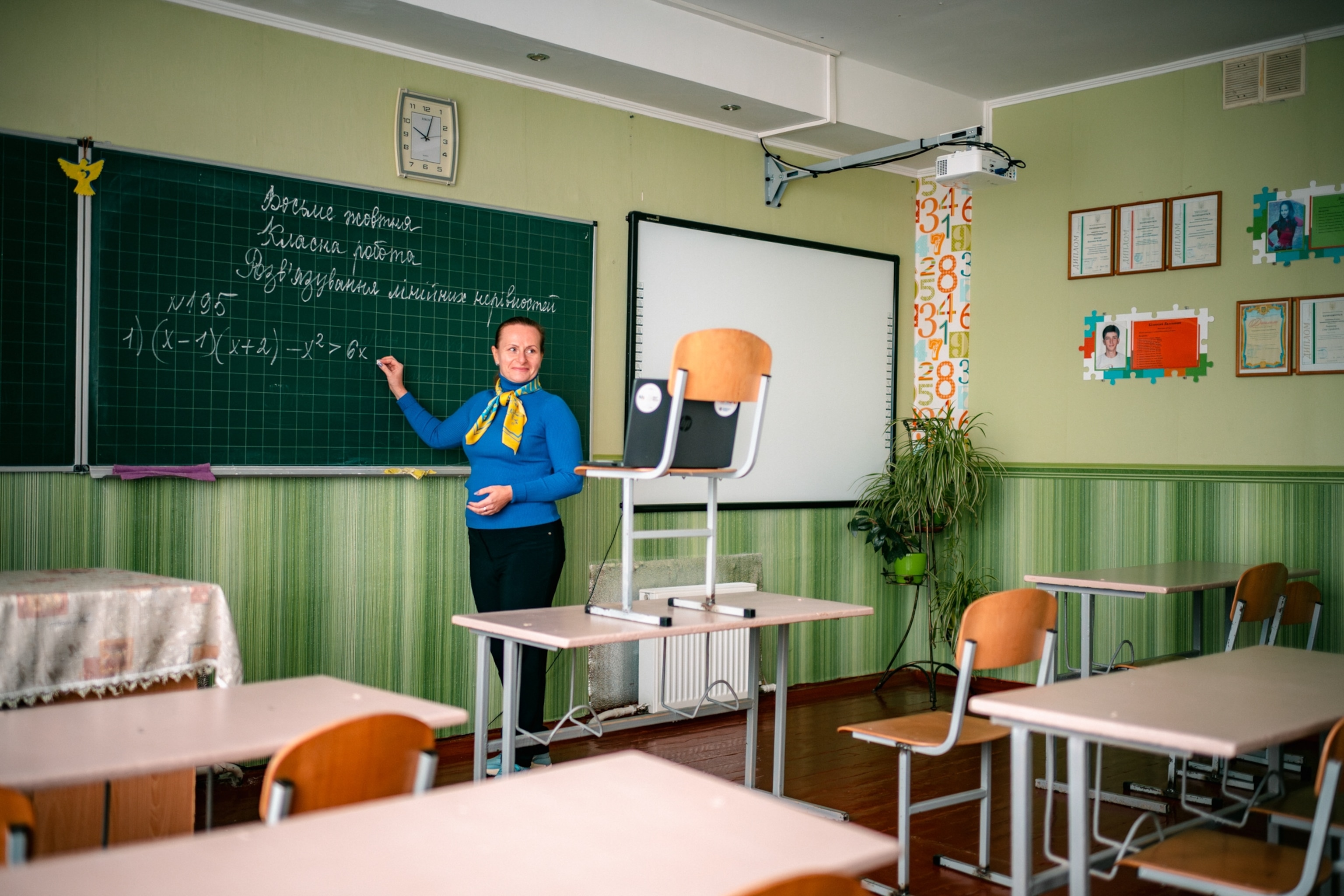 A woman wearing blue stands in front of a chalkboard in a green classroom.