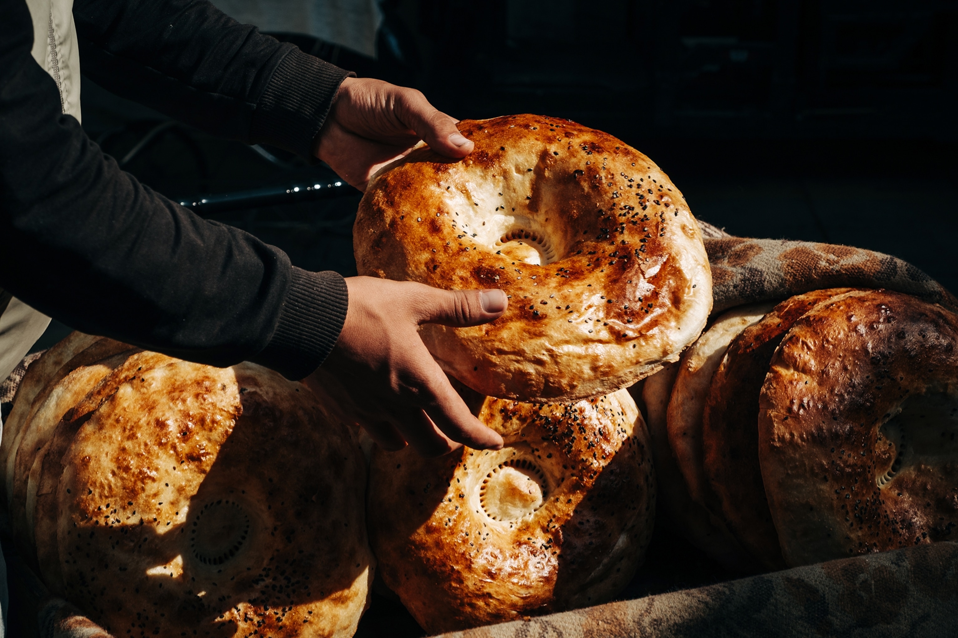 A pair of hands holding up a big, donut-like pastry with a flaky and sesame-sprinkled crust into the sunlight .