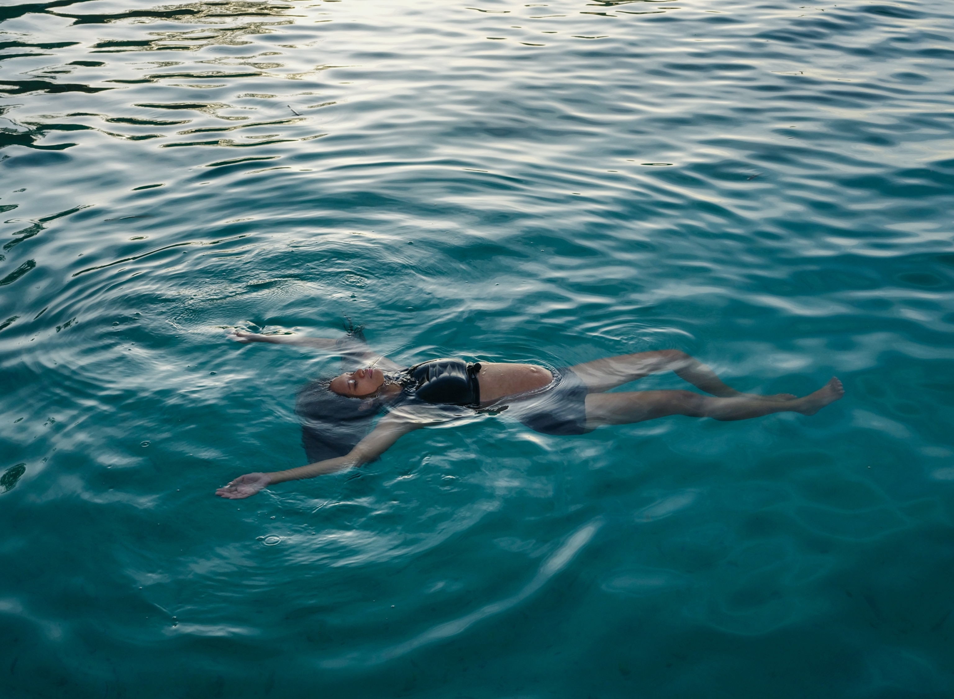 A woman flats on her back in a large, natural body of water. Her belly and face peak above the water while the rest of her is submerged.