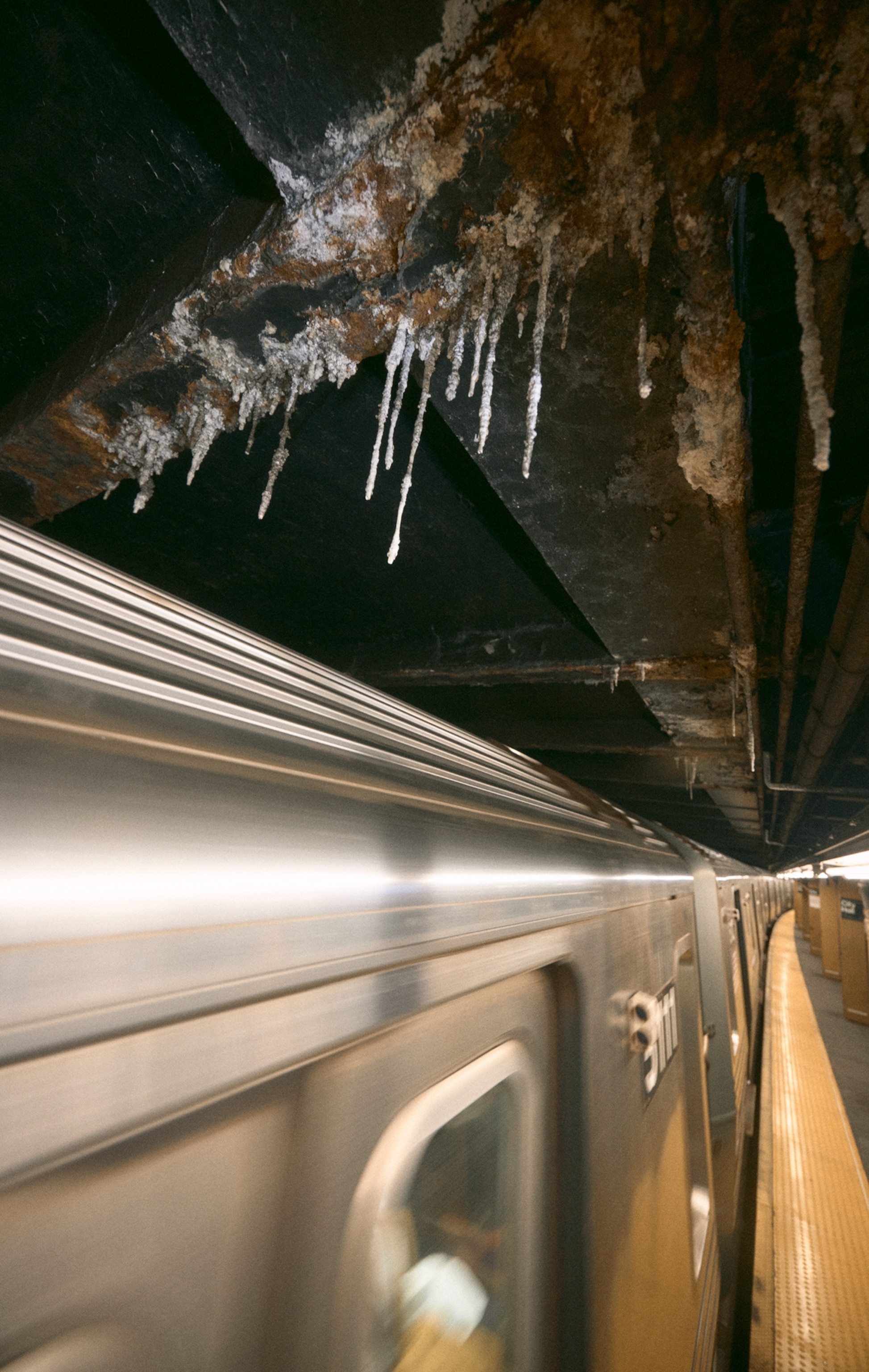 Hanging formations called calthemites hang off the roof of an NYC subway as a train passes by.