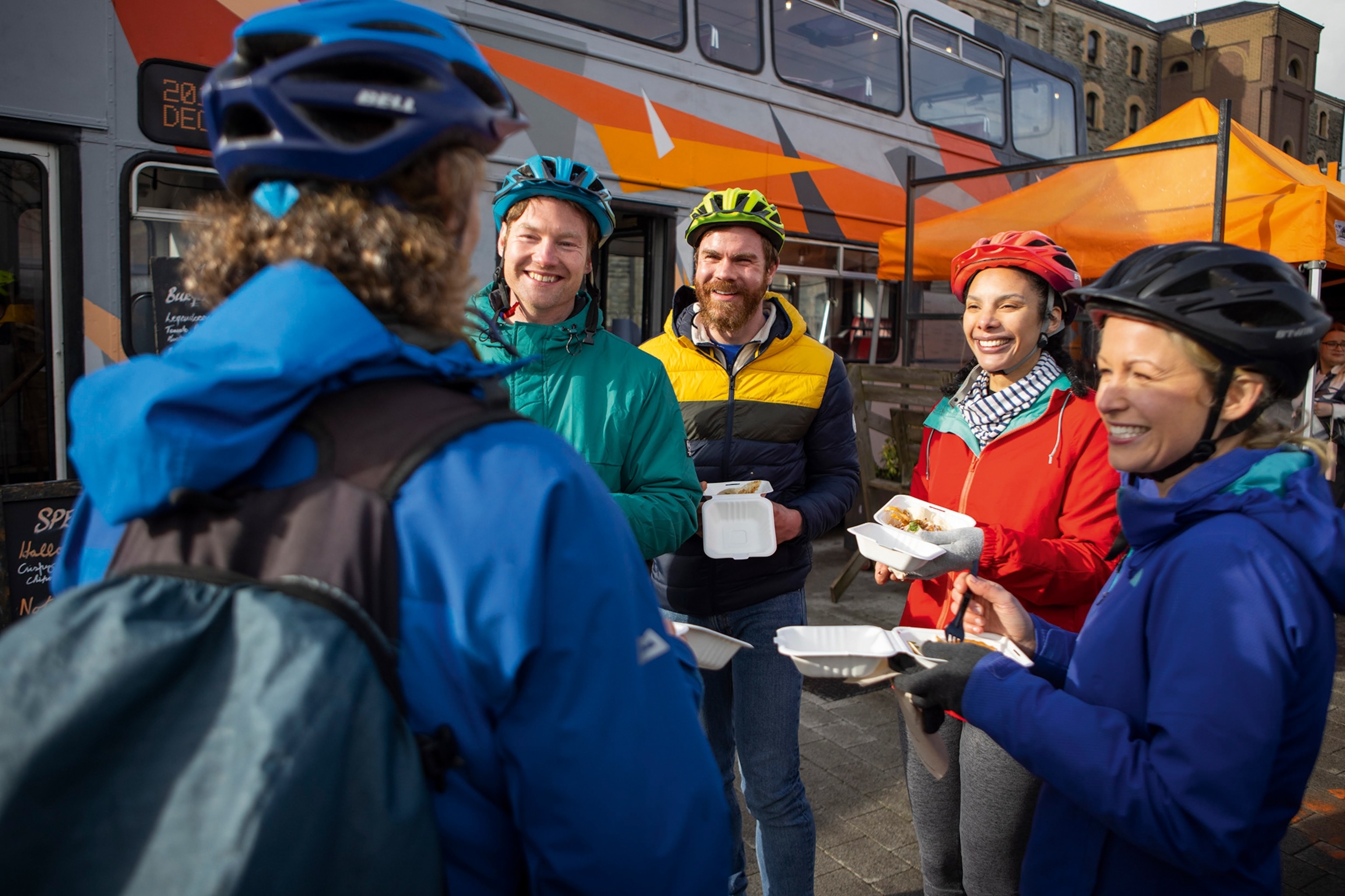 a group of cyclists in windbreakers and helmets stand together and share a bite to eat