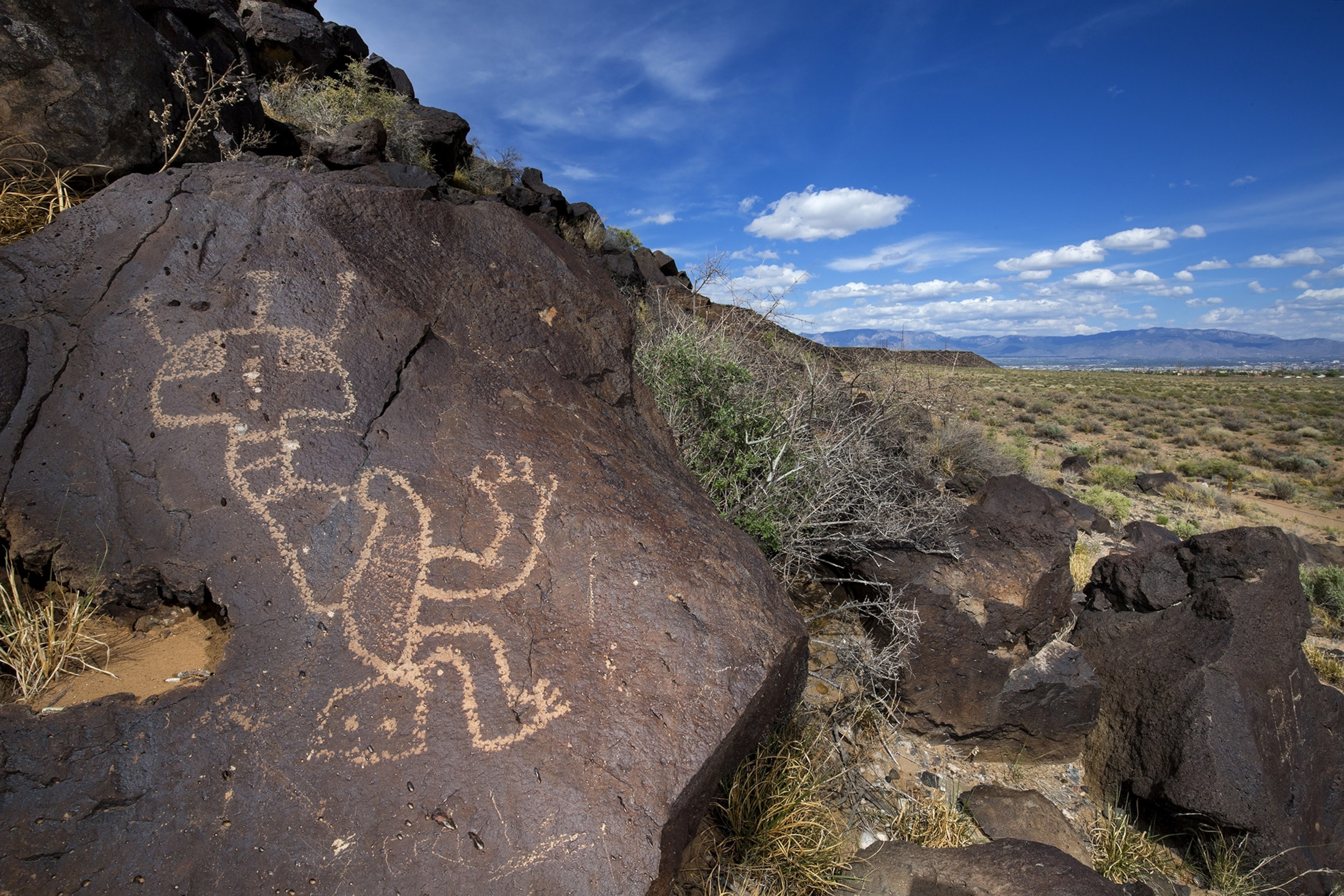 A close-up of a petroglyph in the foreground with rocky desert landscape stretching out behind, blue sky with white clouds above